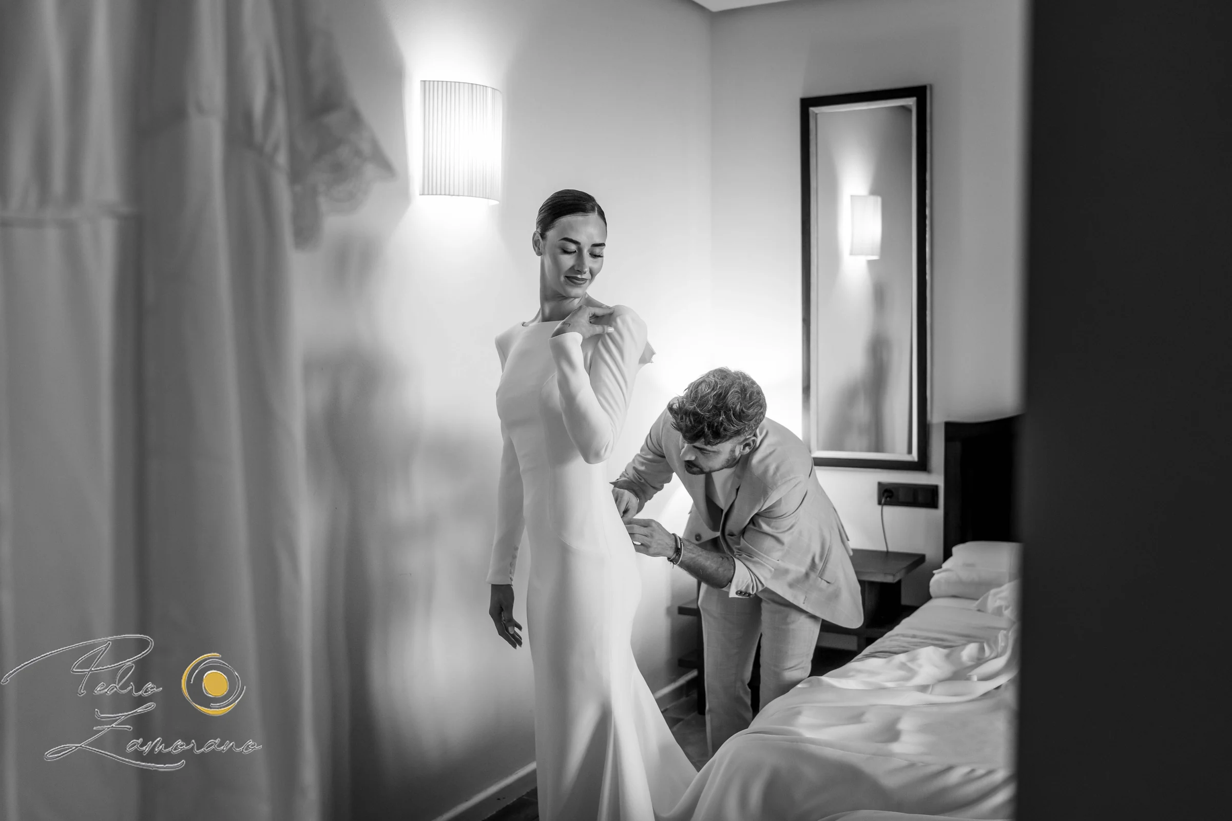 Novia vistiéndose acompañada durante los preparativos en una fotografía en blanco y negro.