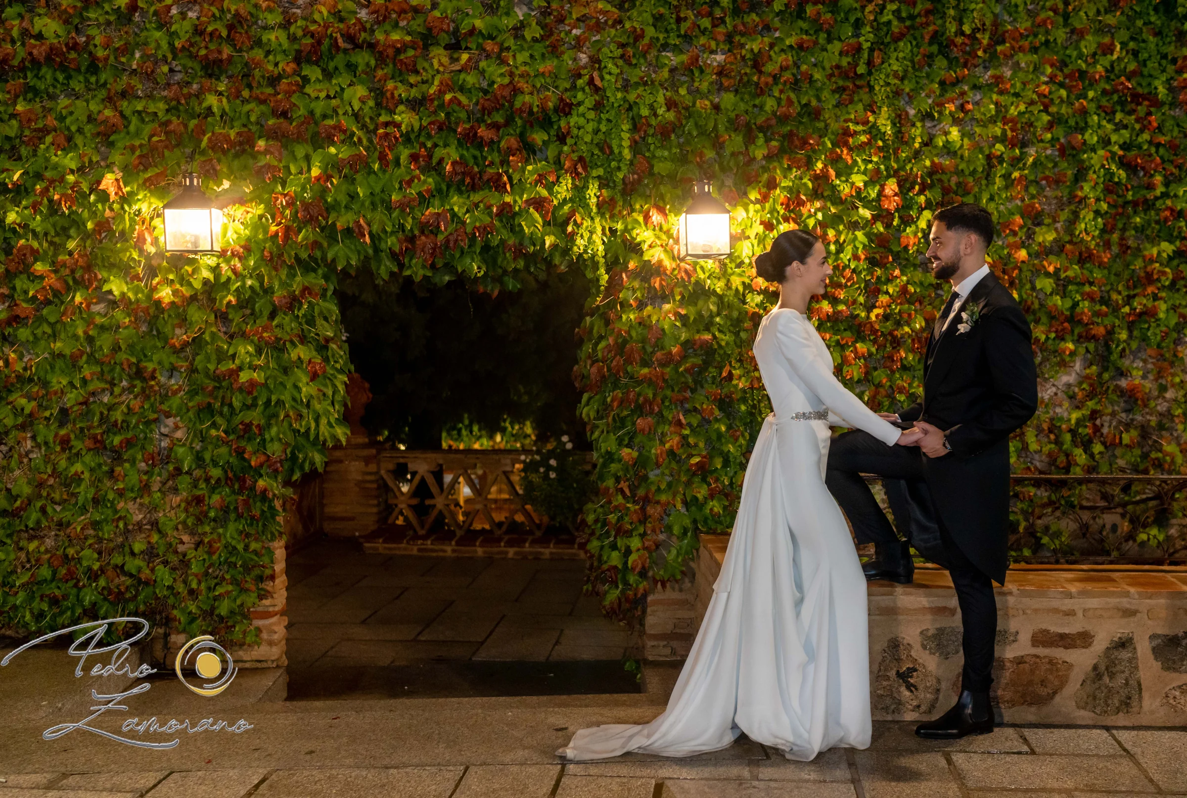 Novios de noche frente a un muro cubierto de hiedra en los jardines.