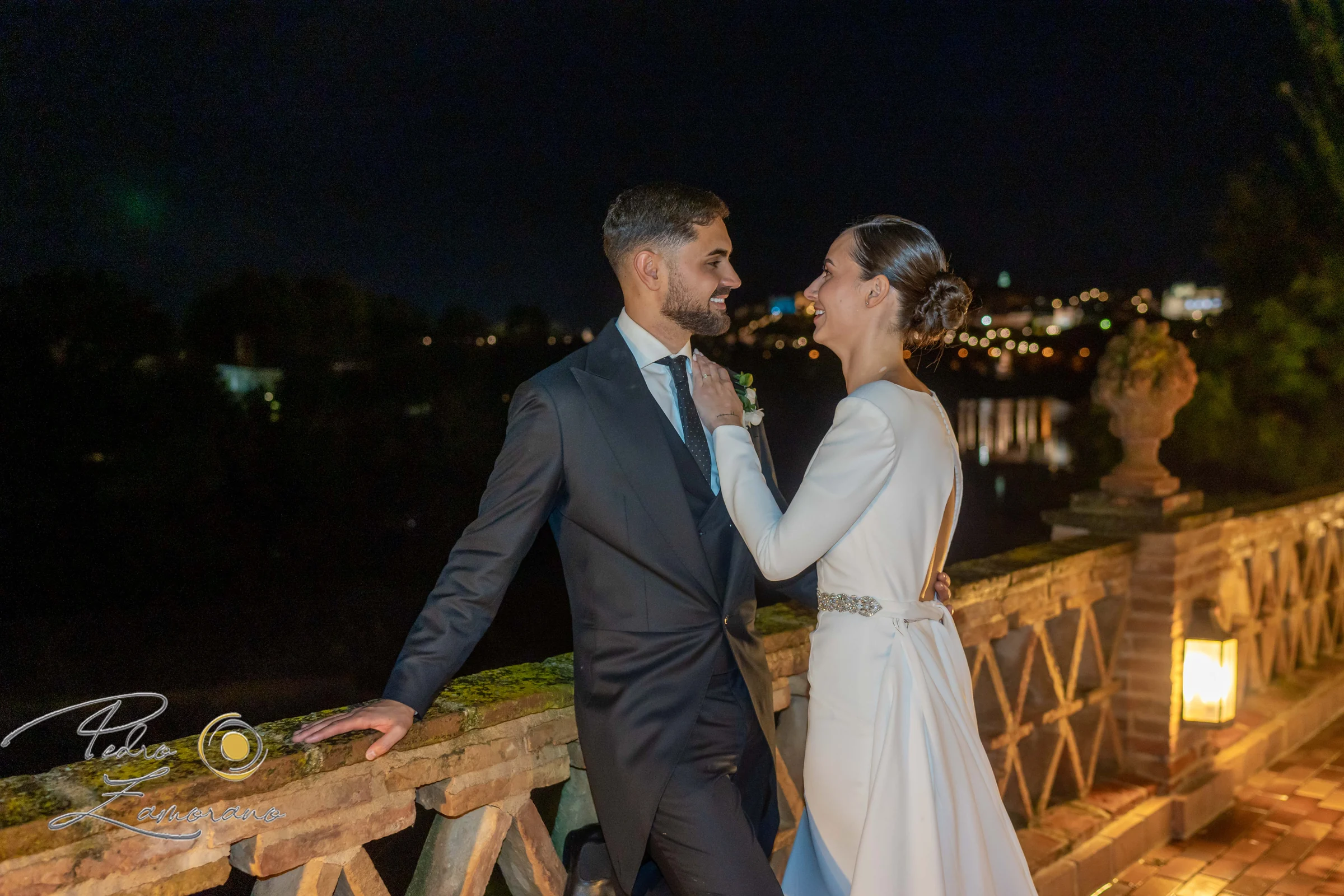 Novios abrazados junto a una terraza iluminada durante la noche.
