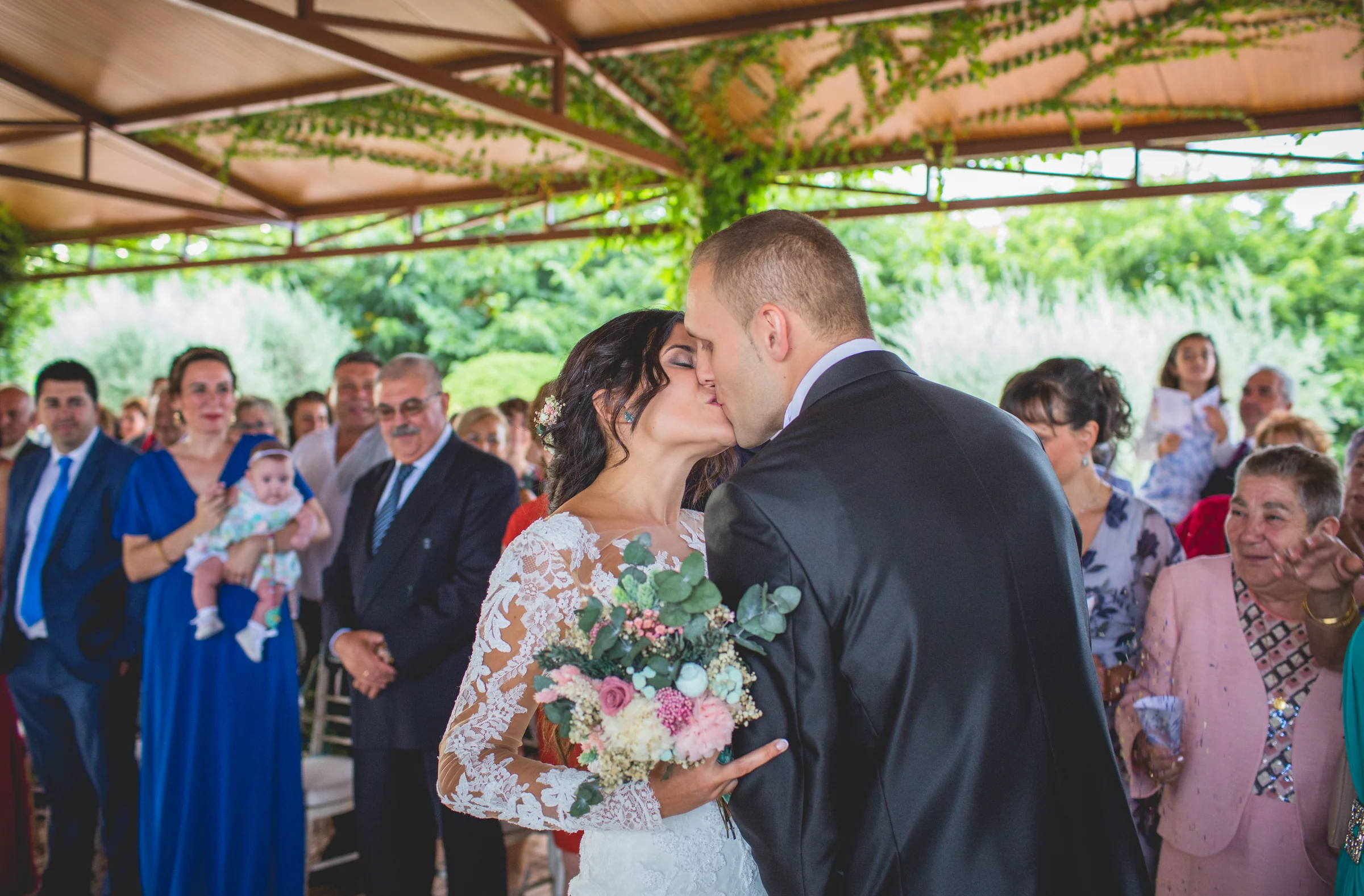 Novios besándose durante la ceremonia rodeados por familiares e invitados.