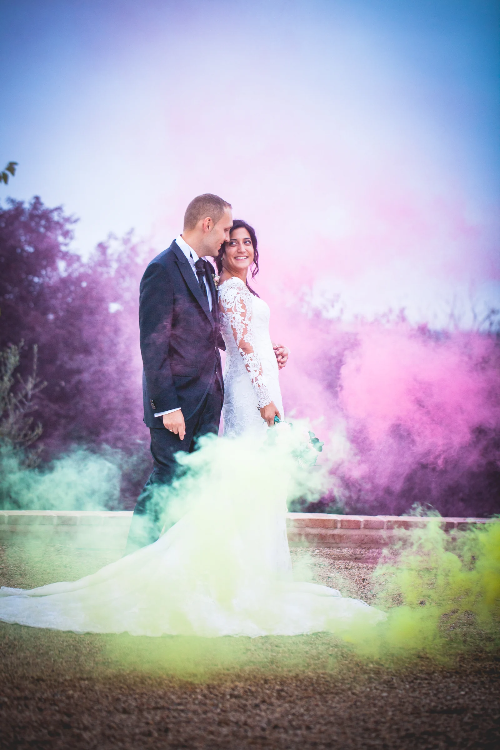 Novios rodeados por humo de colores durante una fotografía creativa en los jardines.