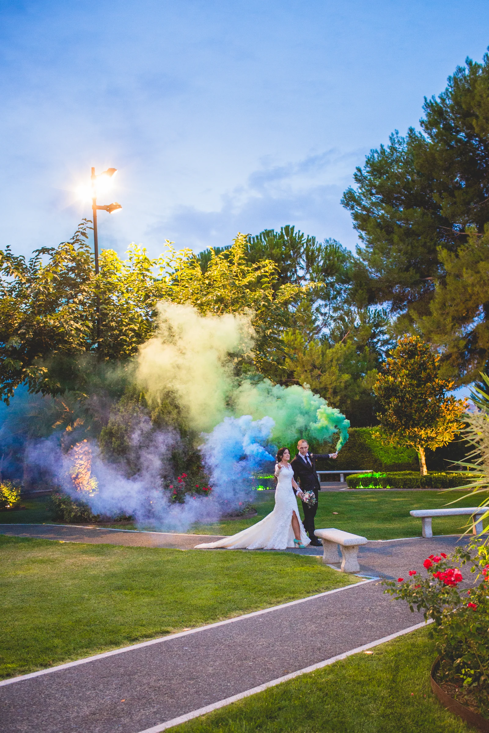 Novios fotografiados a distancia entre humo de colores en los jardines.