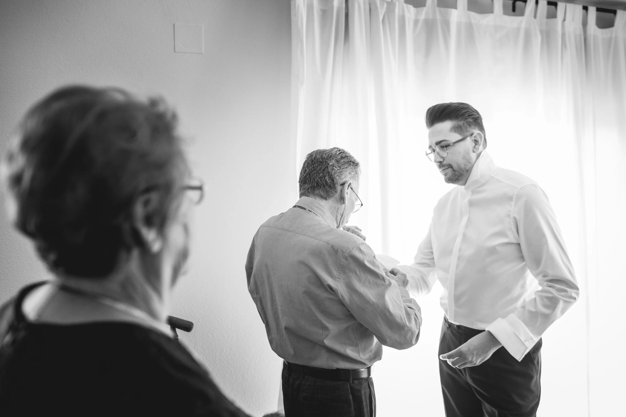Familiares ayudando al novio a vestirse junto a una ventana antes de la ceremonia.