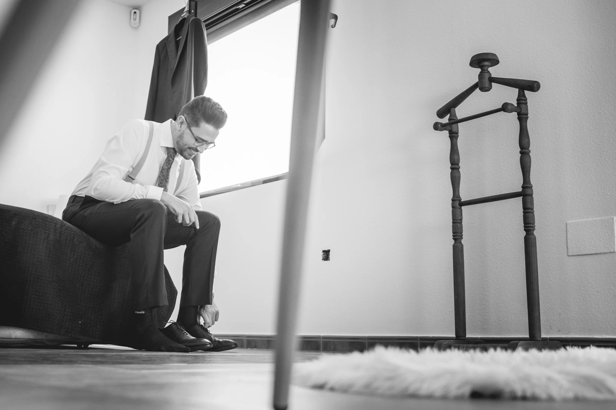 Novio sentado junto a una ventana atándose los zapatos en una fotografía en blanco y negro.