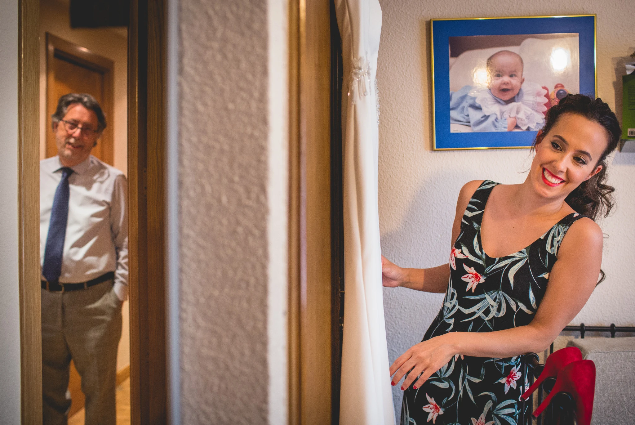 Mujer sonriendo junto al vestido de novia mientras un familiar observa desde la puerta.