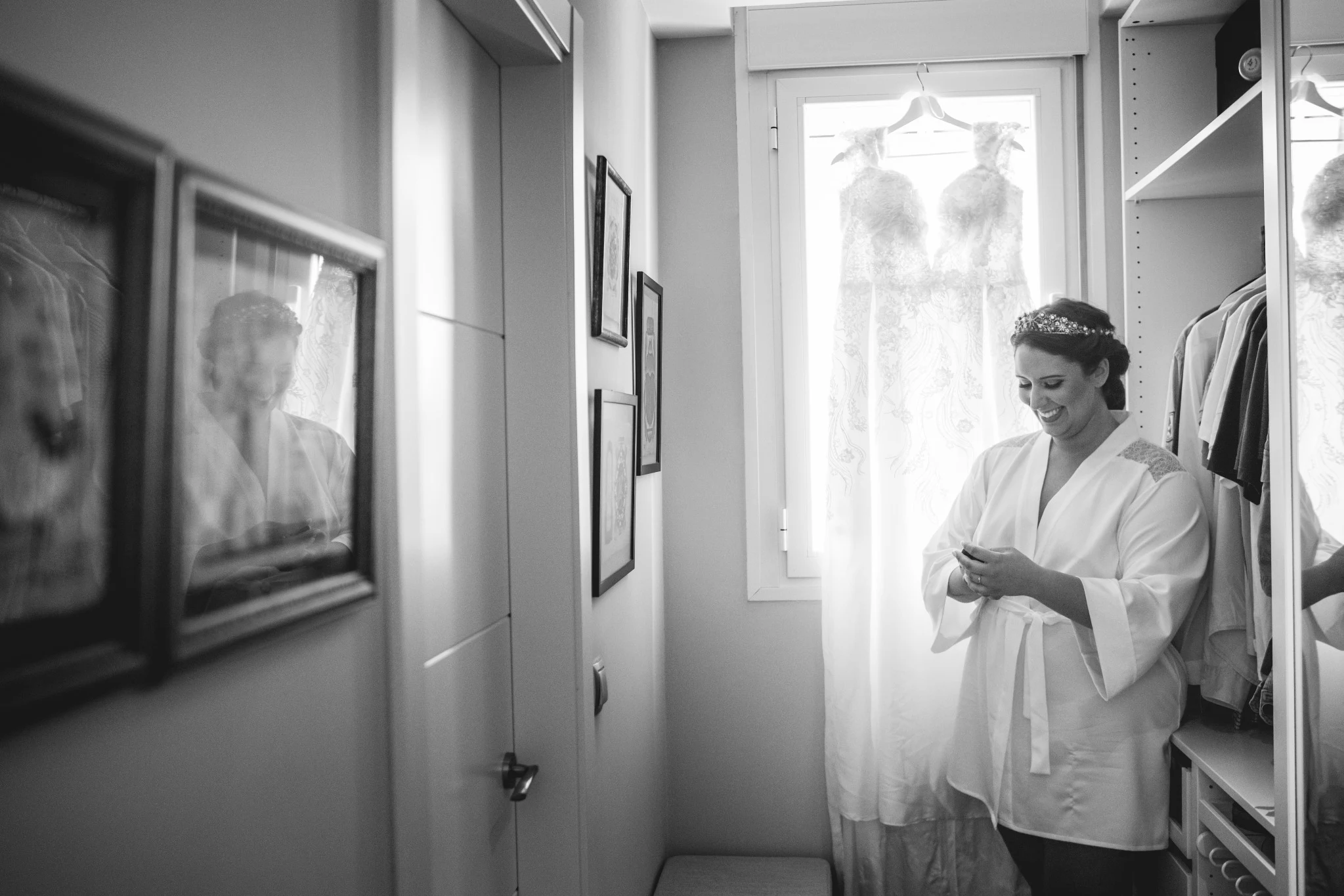 Novia con bata sonriendo mientras se prepara con el vestido de boda colgado junto a la ventana.