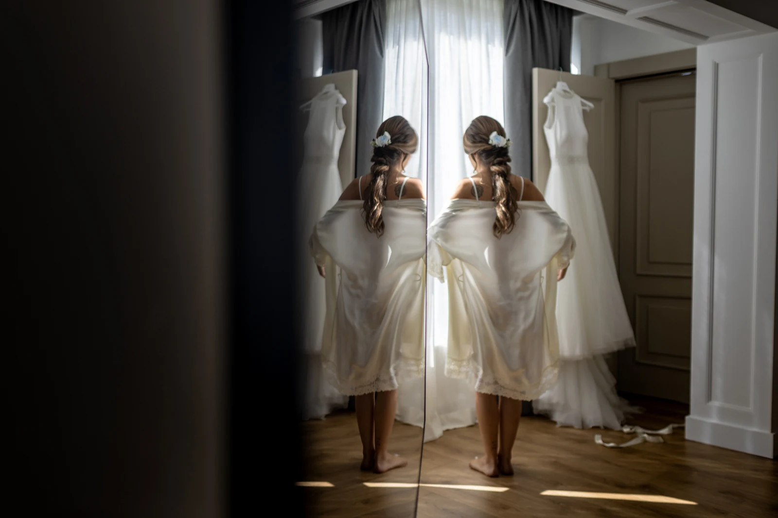 Novia de espaldas ante un espejo mientras su vestido cuelga junto a la ventana.