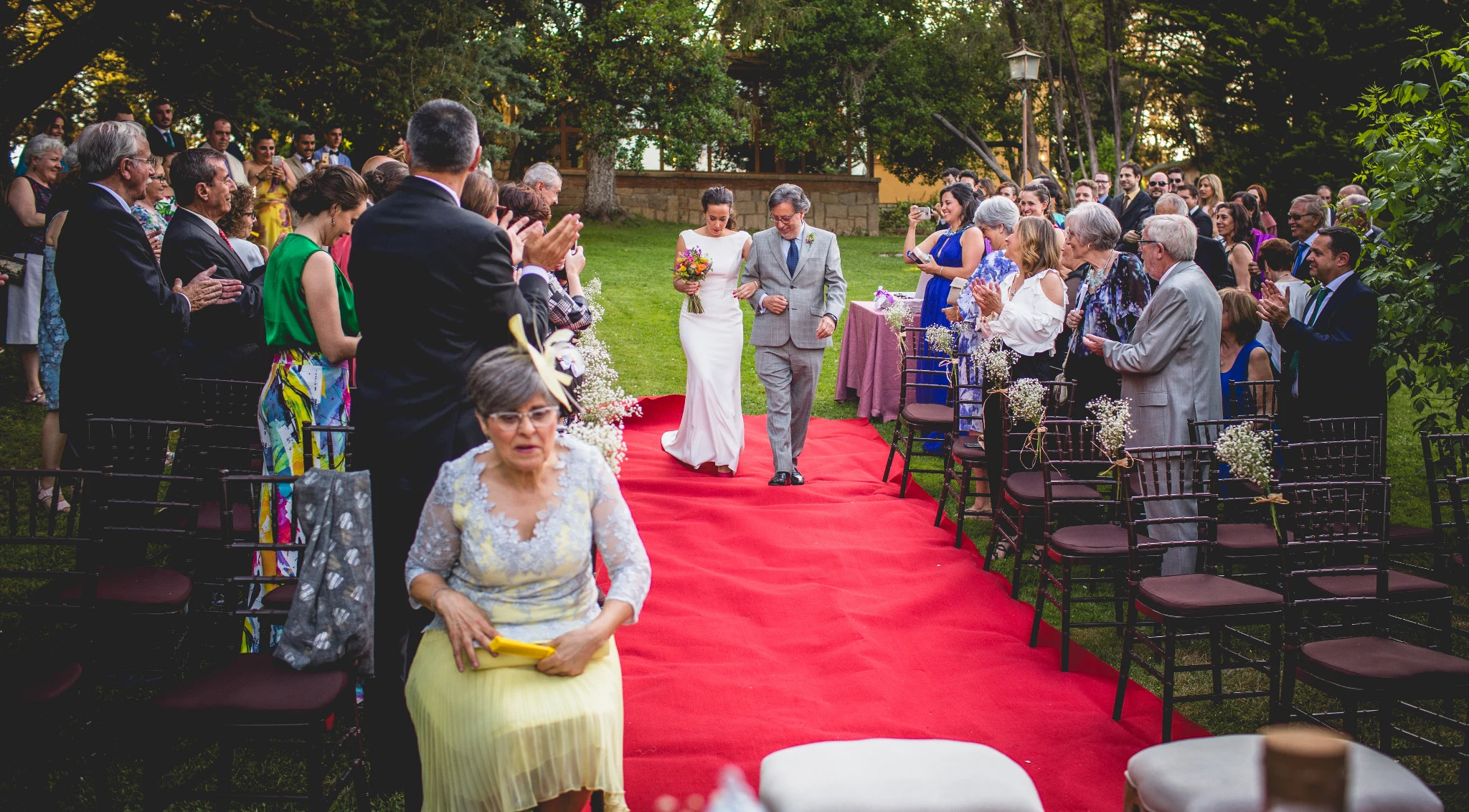 Novia entrando del brazo de su padre por una alfombra roja entre los invitados.