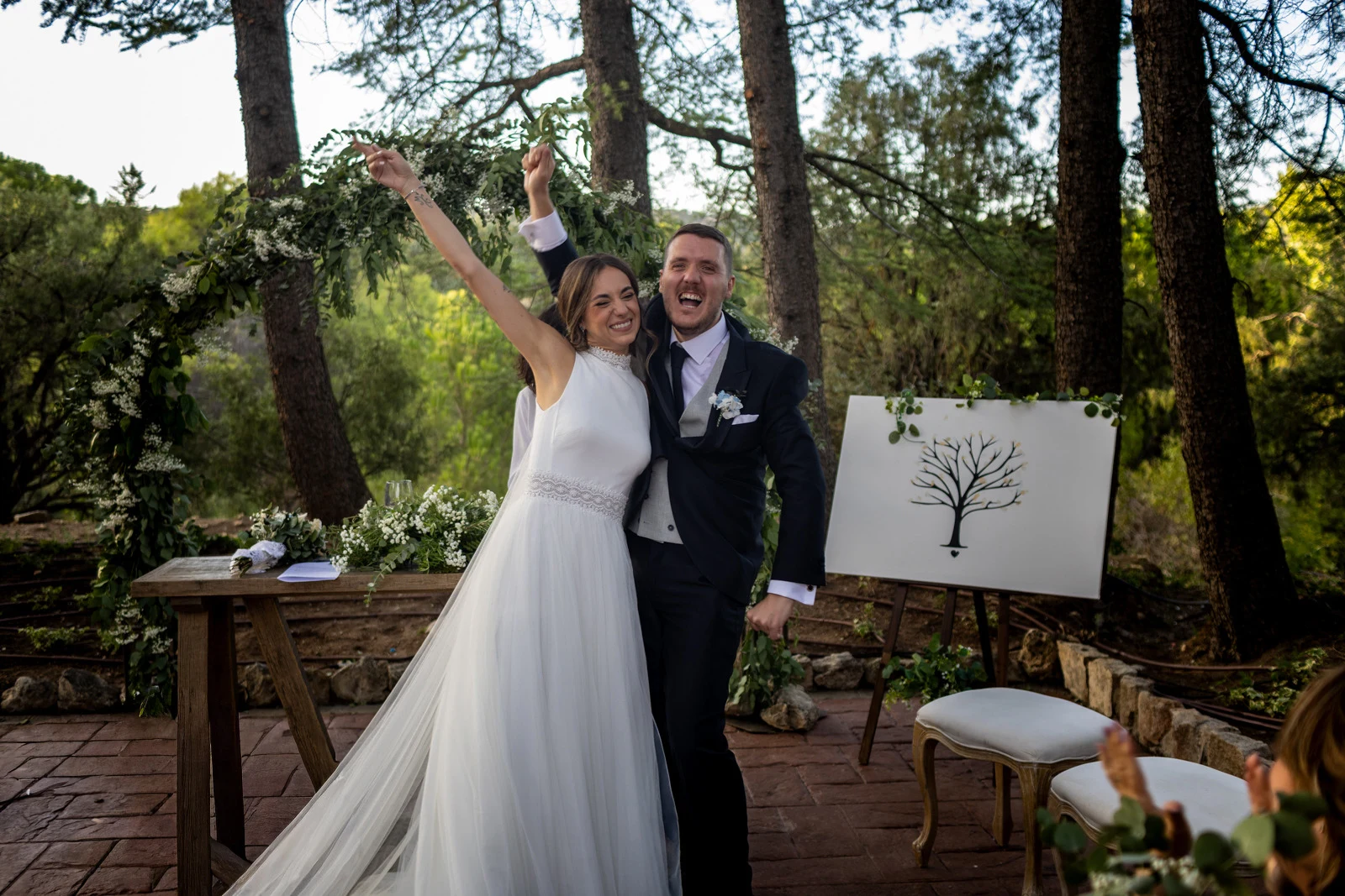 Novios levantan los brazos sonrientes después de la ceremonia bajo un arco vegetal.