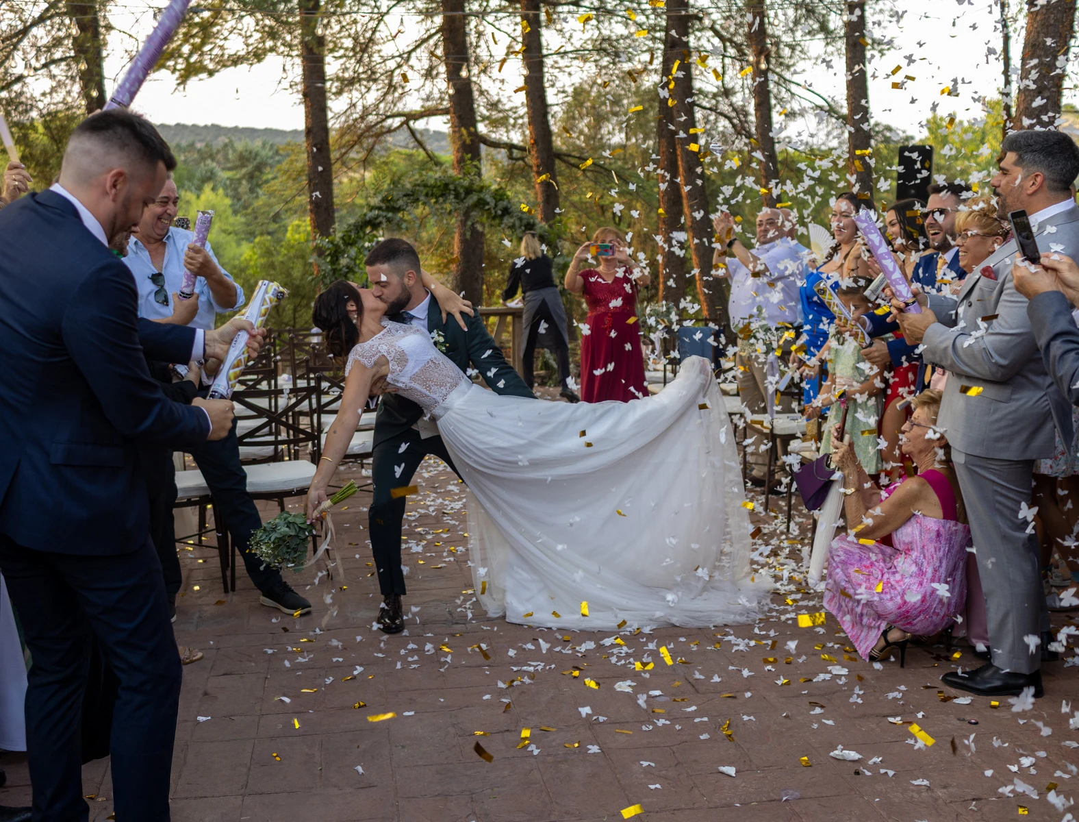 Novio inclina a la novia para besarla mientras los invitados lanzan confeti.