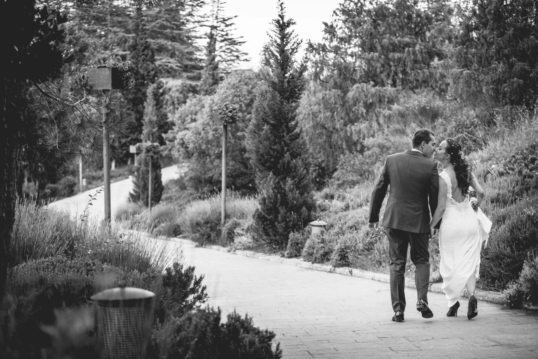 Novios paseando de la mano por un camino ajardinado mientras se besan.