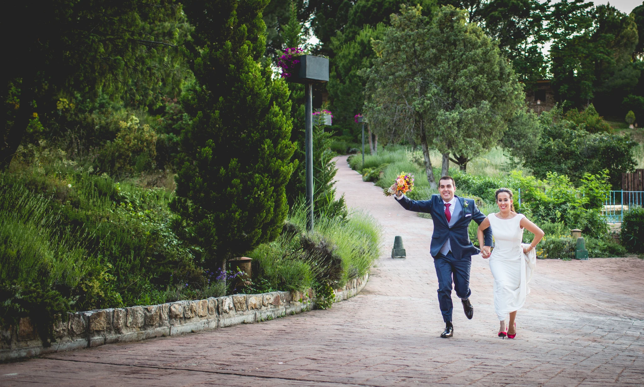 Novios corren de la mano por un camino rodeado de vegetación y árboles.