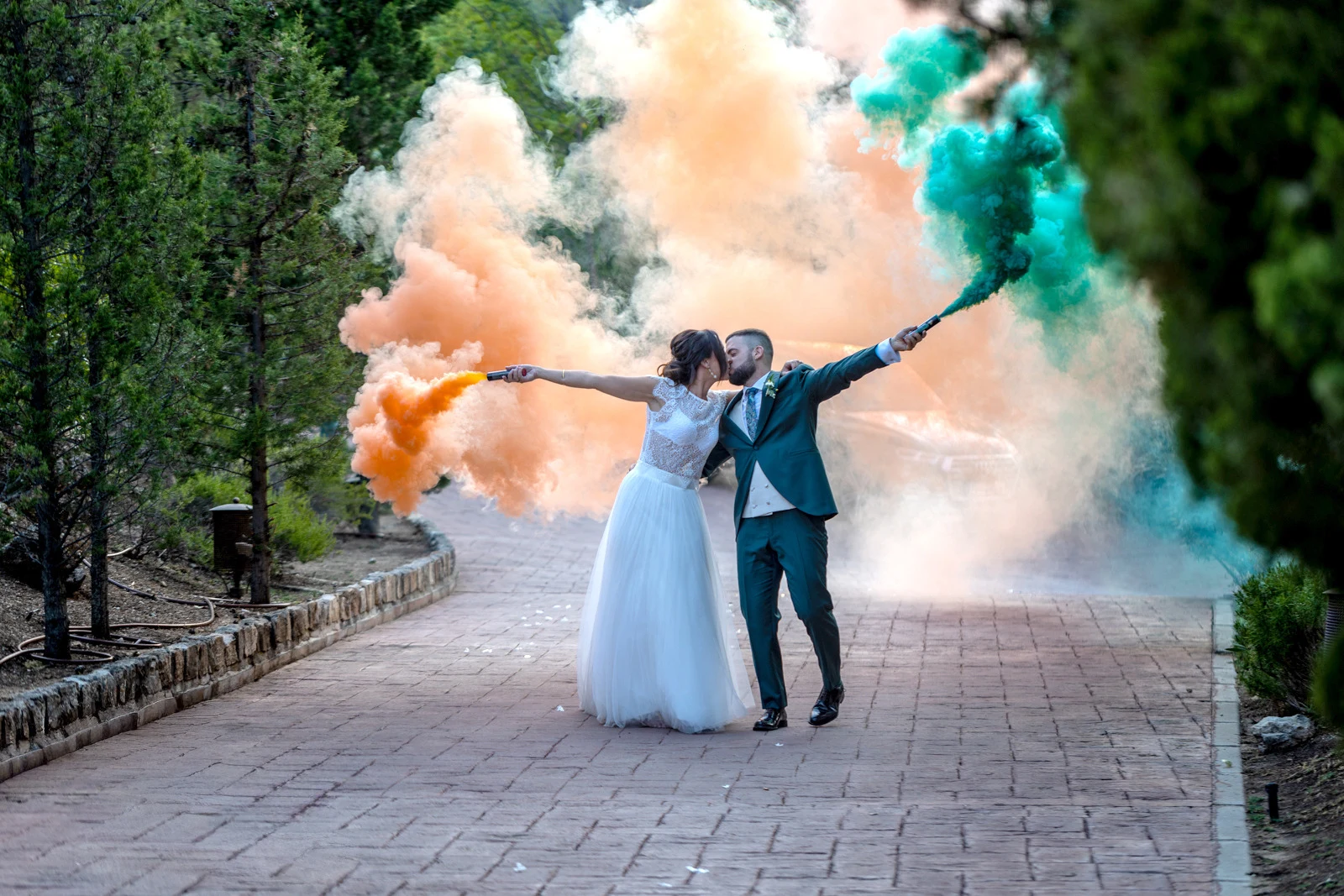 Novios besándose mientras sostienen bengalas de humo naranja y verde en un camino.