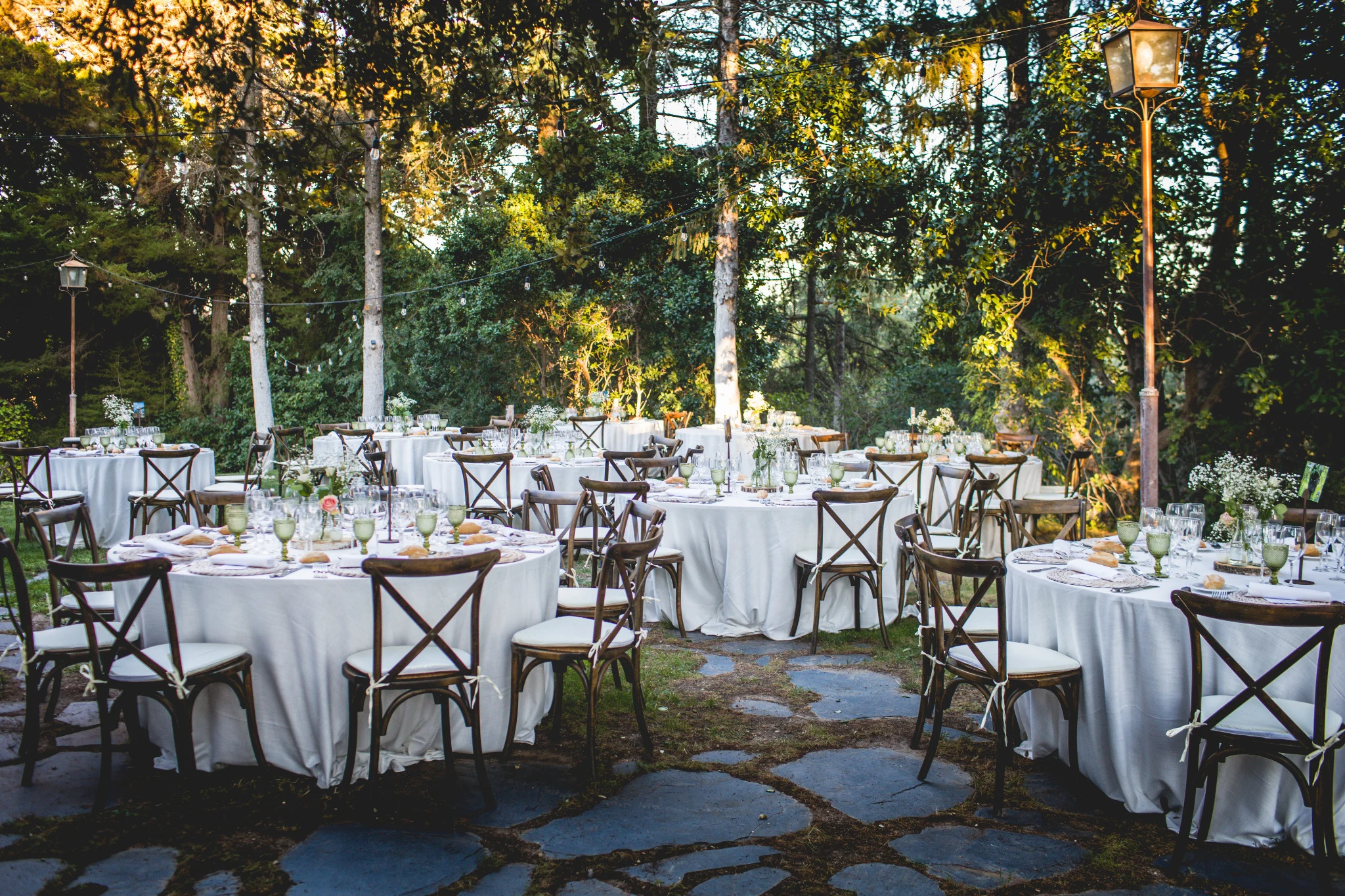 Mesas redondas preparadas para un banquete de boda entre árboles y luces decorativas.