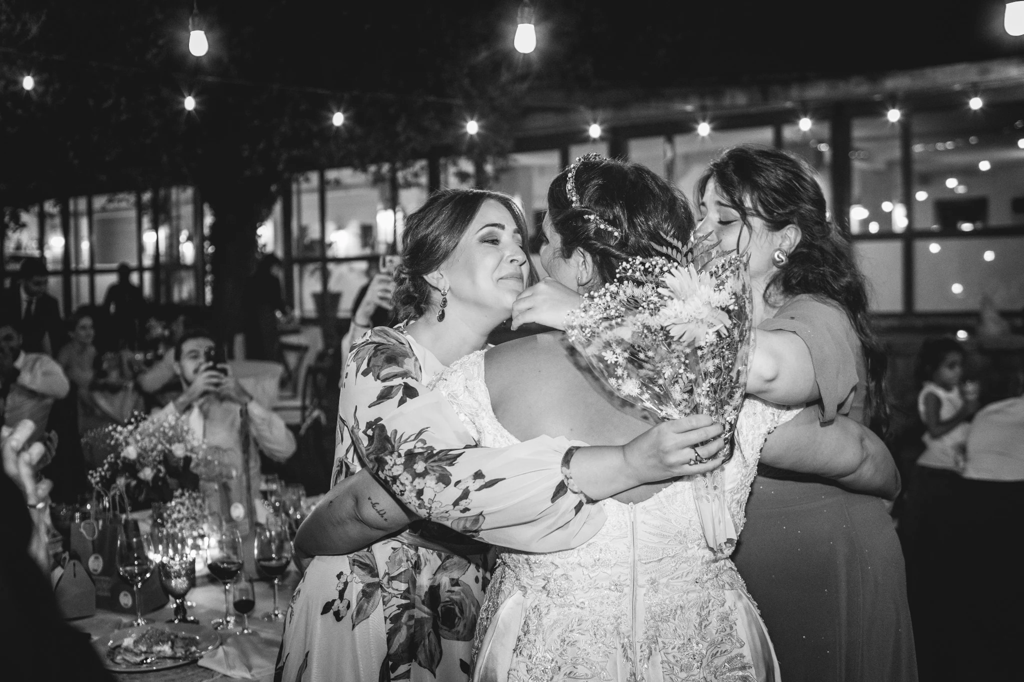 Novia abraza emocionada a dos amigas durante el banquete en una fotografía en blanco y negro.