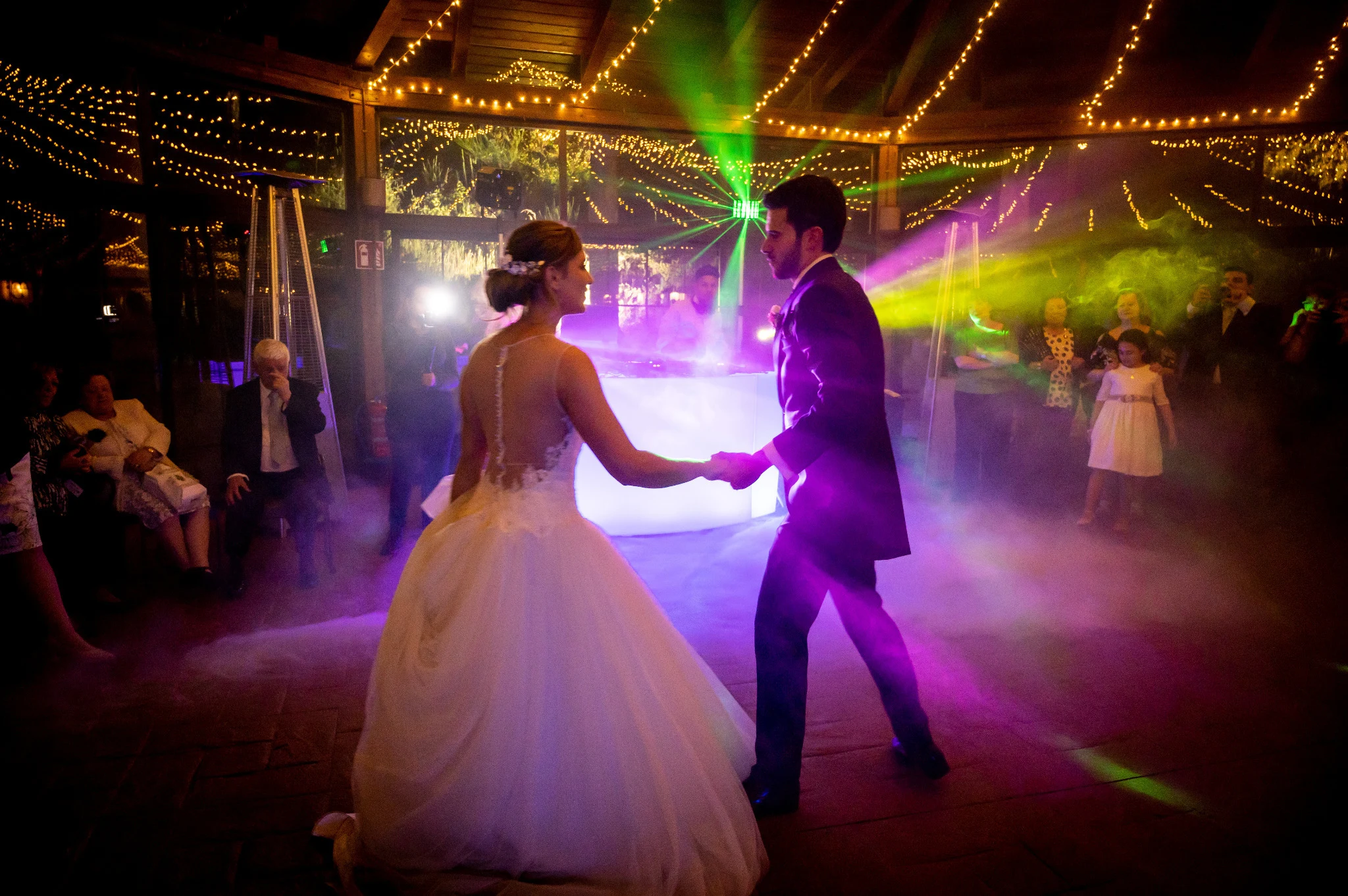 Novios bailan cogidos de la mano entre luces de DJ y humo durante la fiesta.