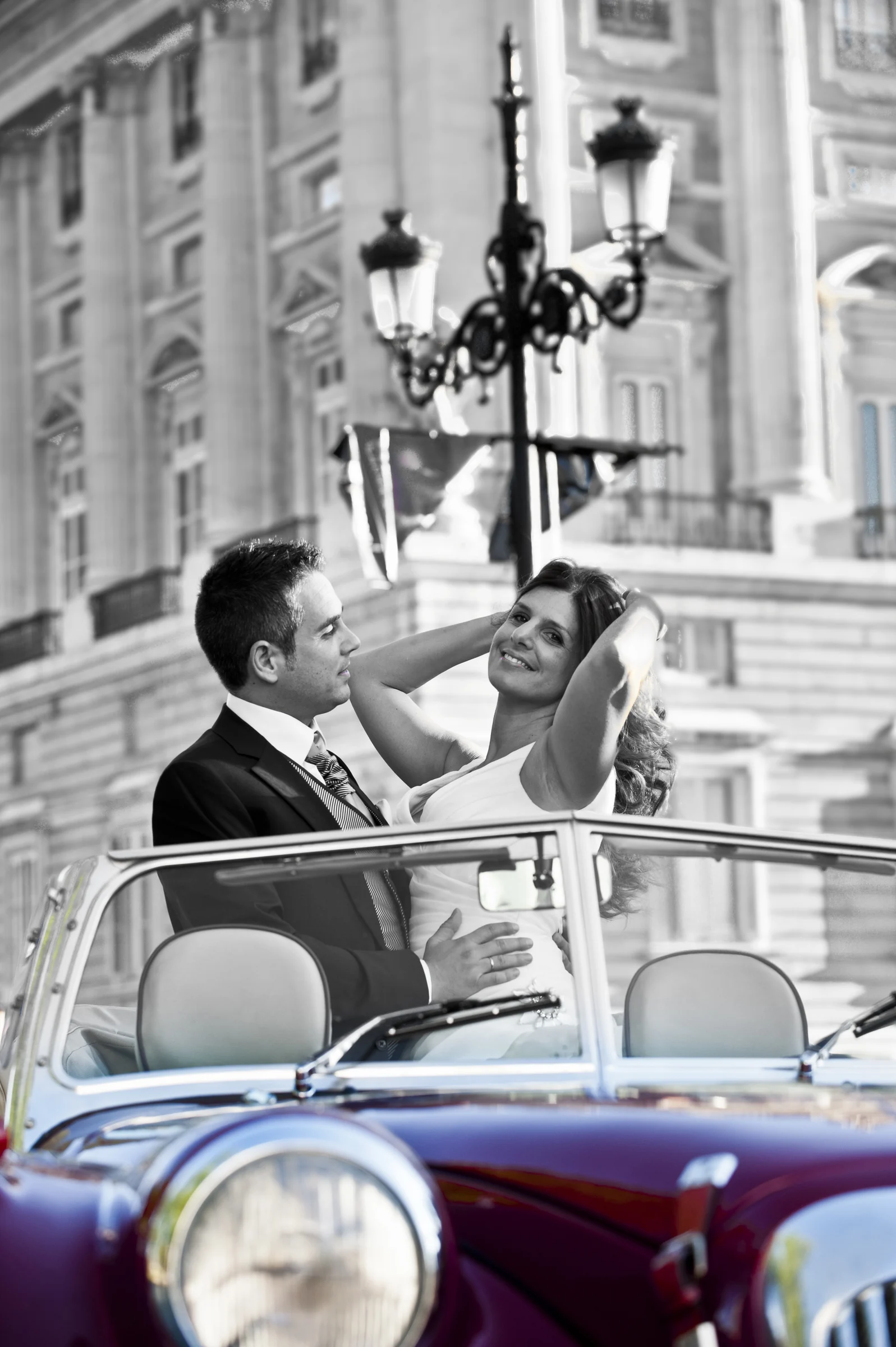 Una pareja de novios posa en el Hurtan descapotable frente al Palacio Real de Madrid en un retrato vertical.