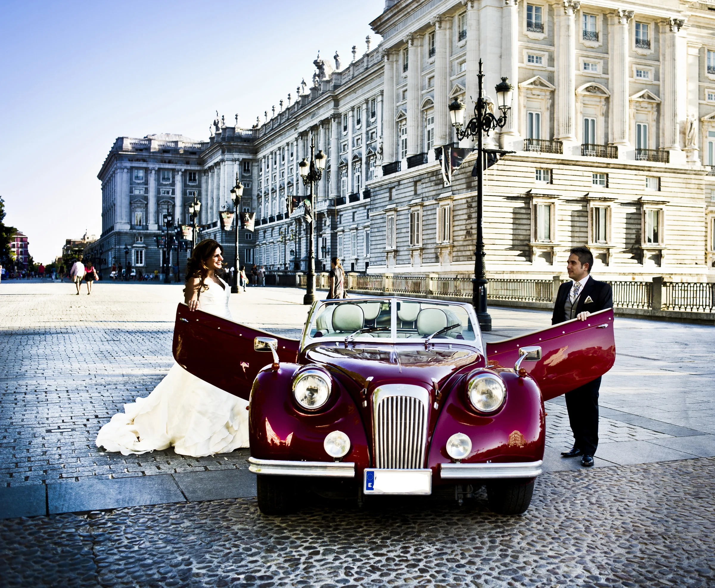 Los novios posan a ambos lados del Hurtan rojo cereza con las puertas abiertas frente al Palacio Real de Madrid.
