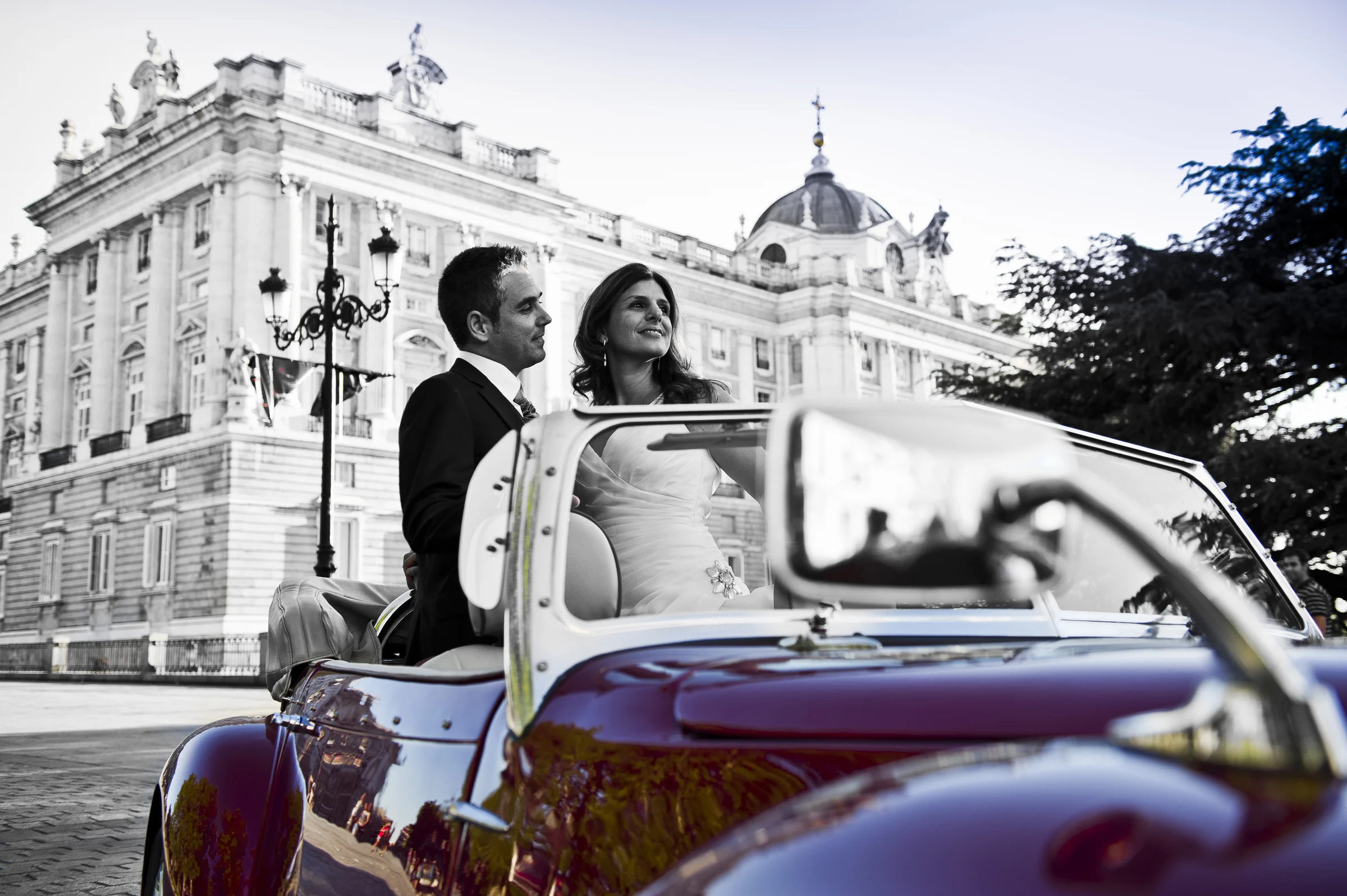 Una pareja de novios posa tras el parabrisas del Hurtan rojo cereza con el Palacio Real de Madrid al fondo.