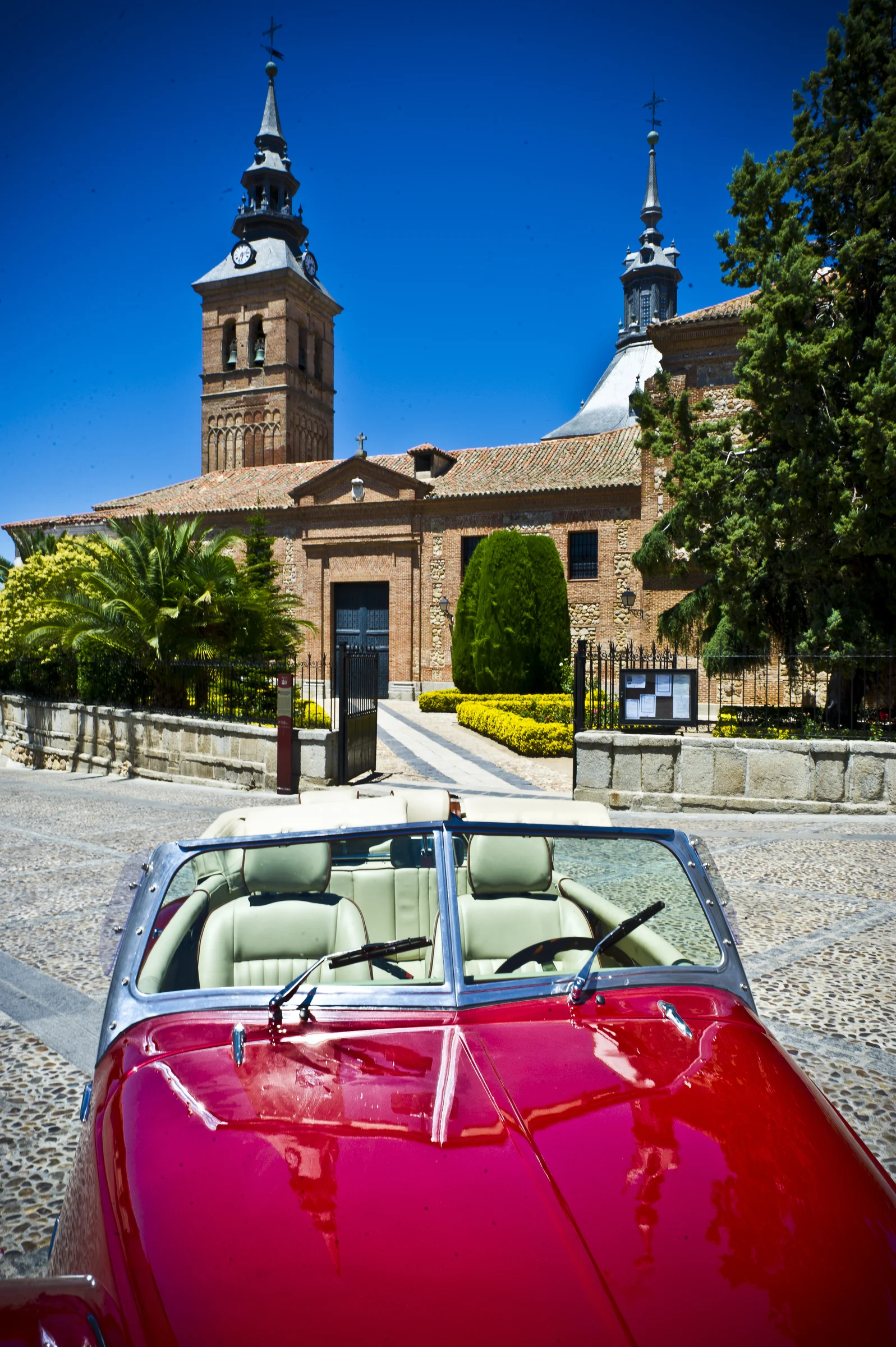Capó rojo cereza del Hurtan en primer plano con una iglesia histórica al fondo.