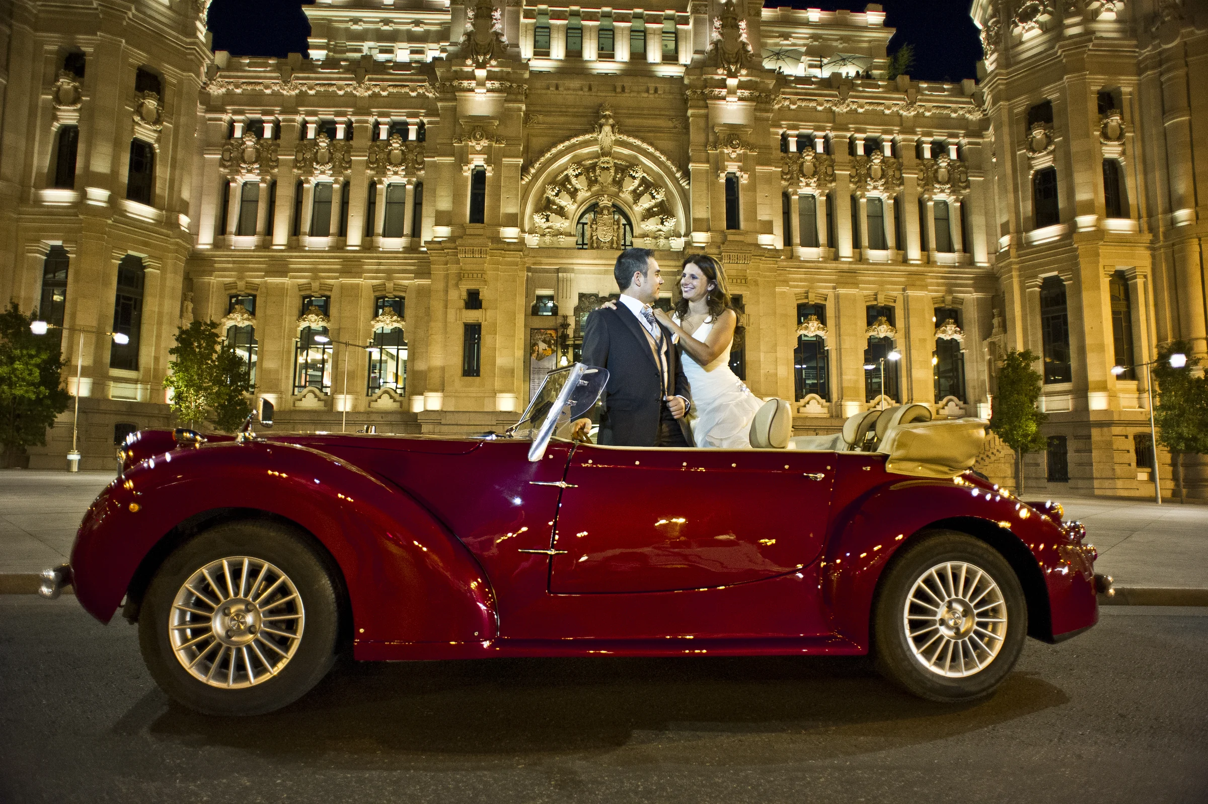 Vista lateral completa del Hurtan rojo cereza con los novios frente al Palacio de Cibeles iluminado por la noche.