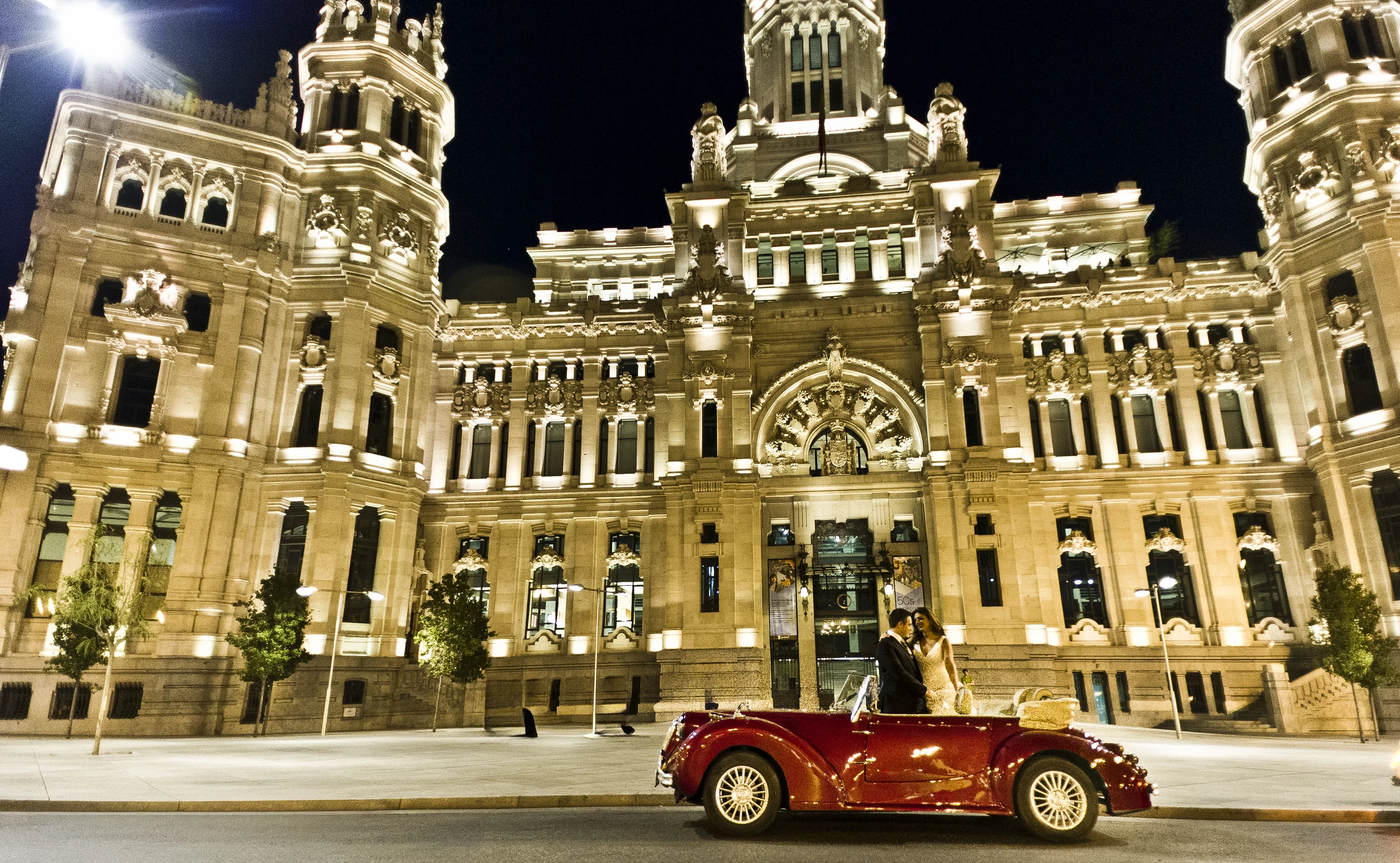 Hurtan rojo cereza con los novios ante el Palacio de Cibeles en una vista nocturna amplia de Madrid.