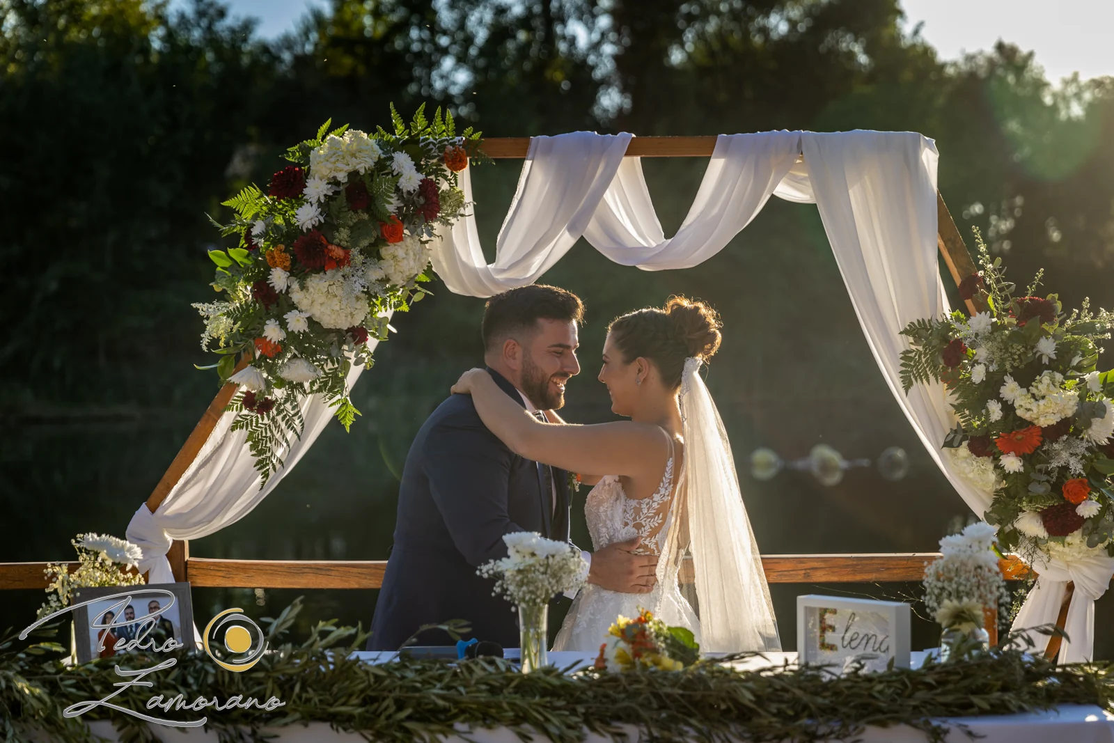 Novios bajo arco floral junto al lago en una boda en Complejo La Cigüeña
