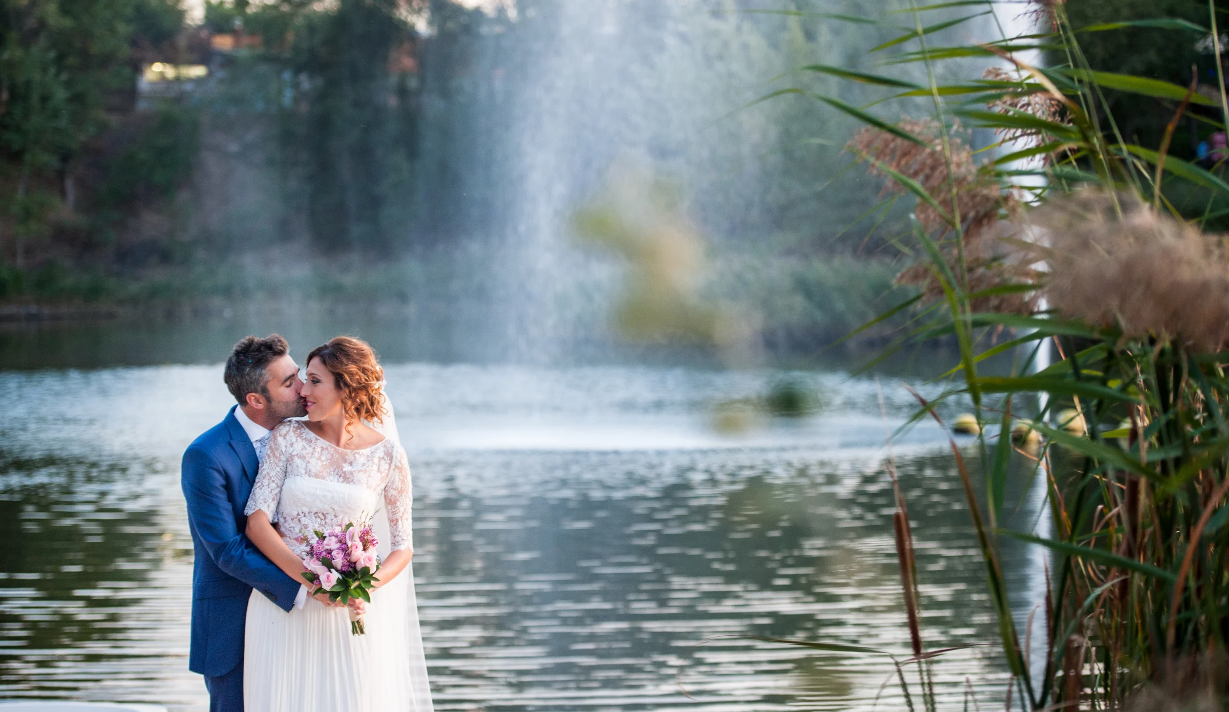 Novios besándose frente a la fuente del lago en una boda en Complejo La Cigüeña