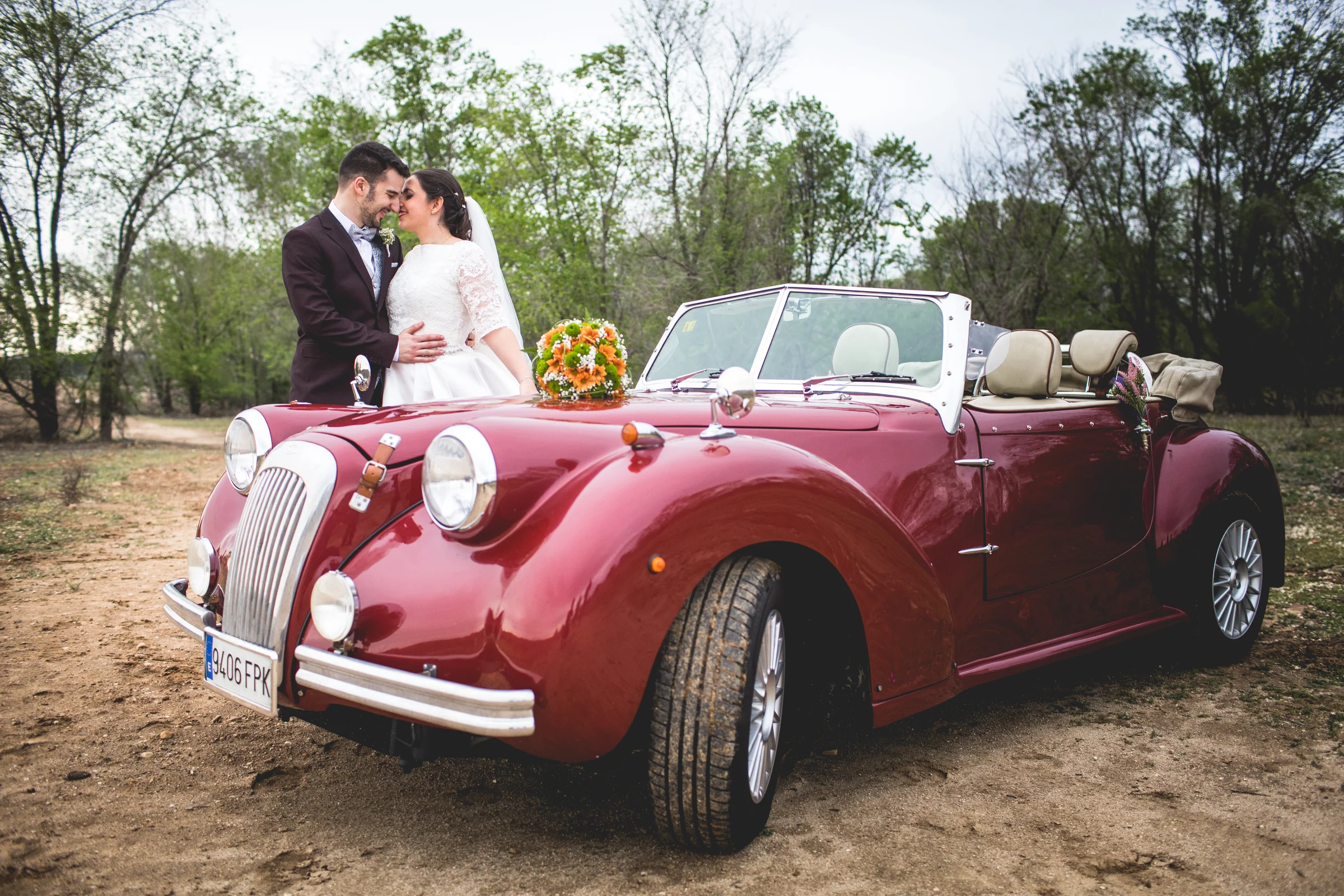Novios posando con coche clásico rojo en una boda en Complejo La Cigüeña