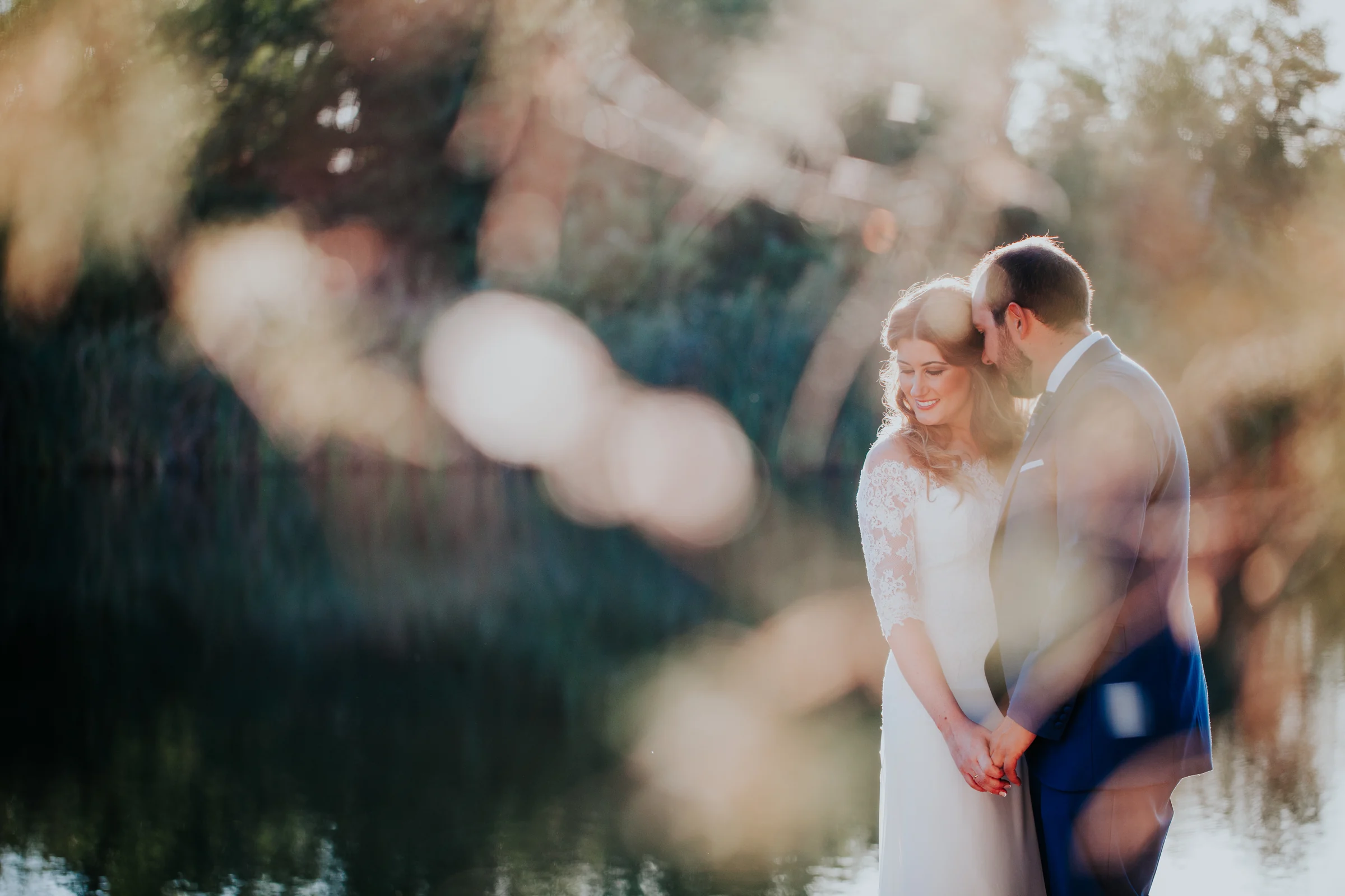 Novios junto al lago entre vegetación desenfocada en una boda en Complejo La Cigüeña
