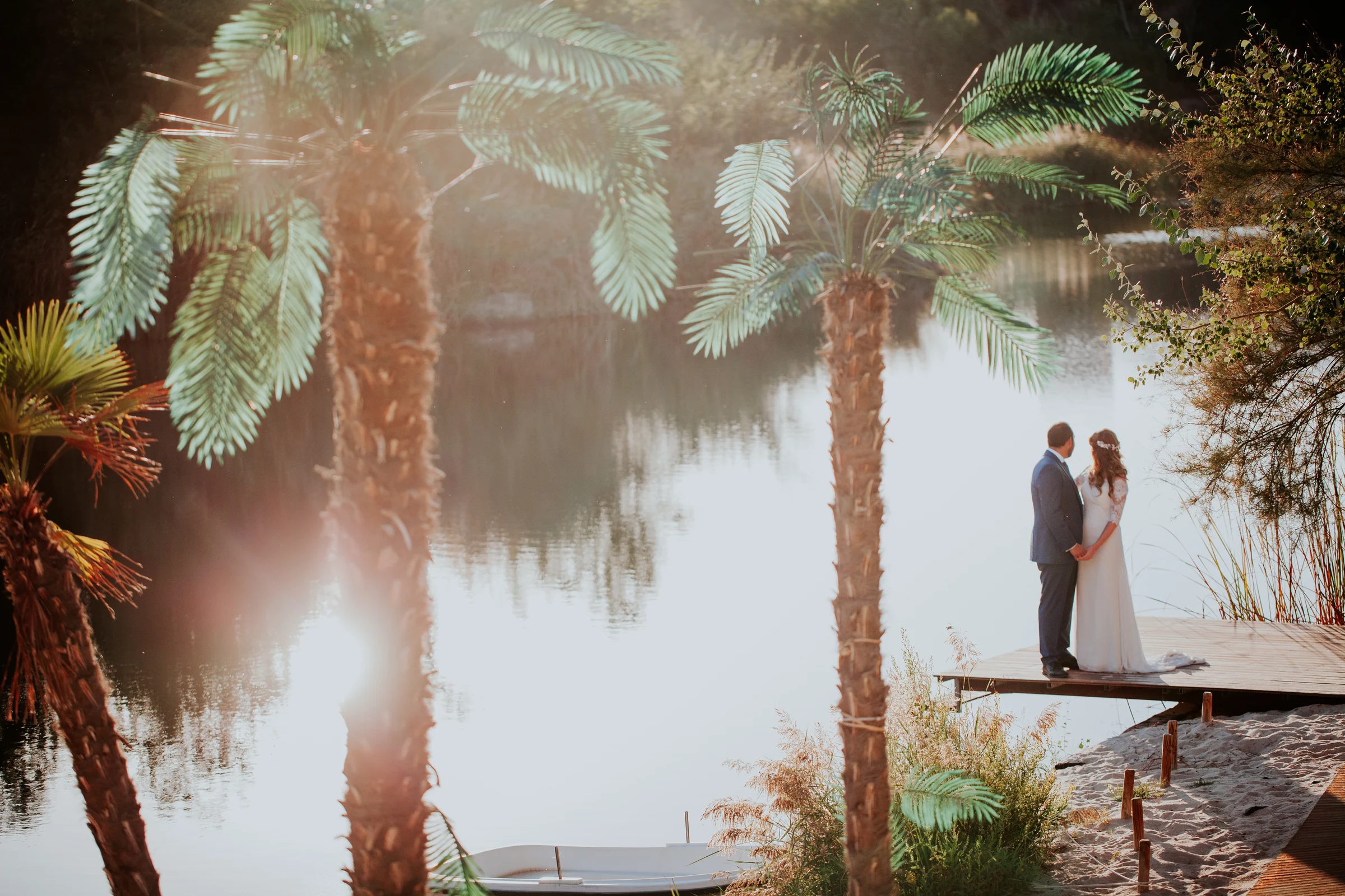 Novios en una pasarela junto a palmeras y lago en una boda en Complejo La Cigüeña