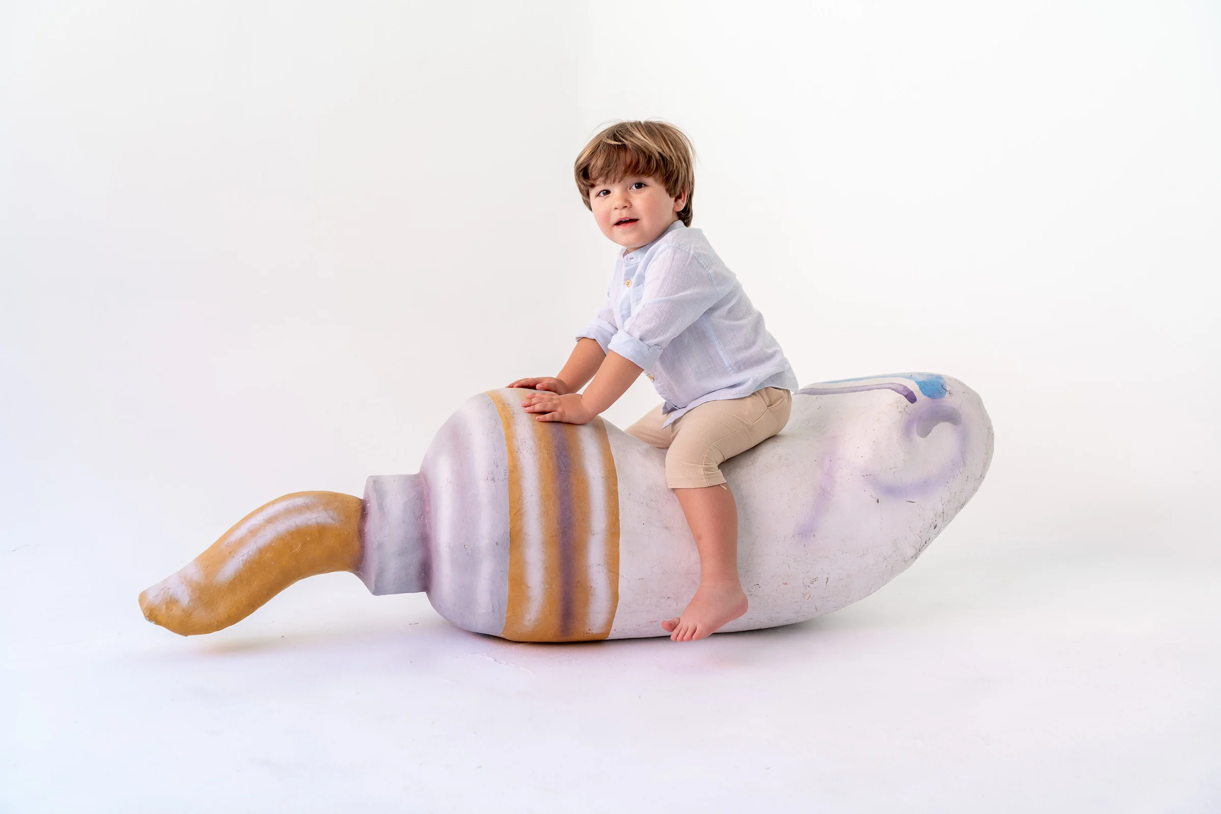 Niño pequeño sentado sobre un atrezo con forma de caracola en fondo blanco de estudio.