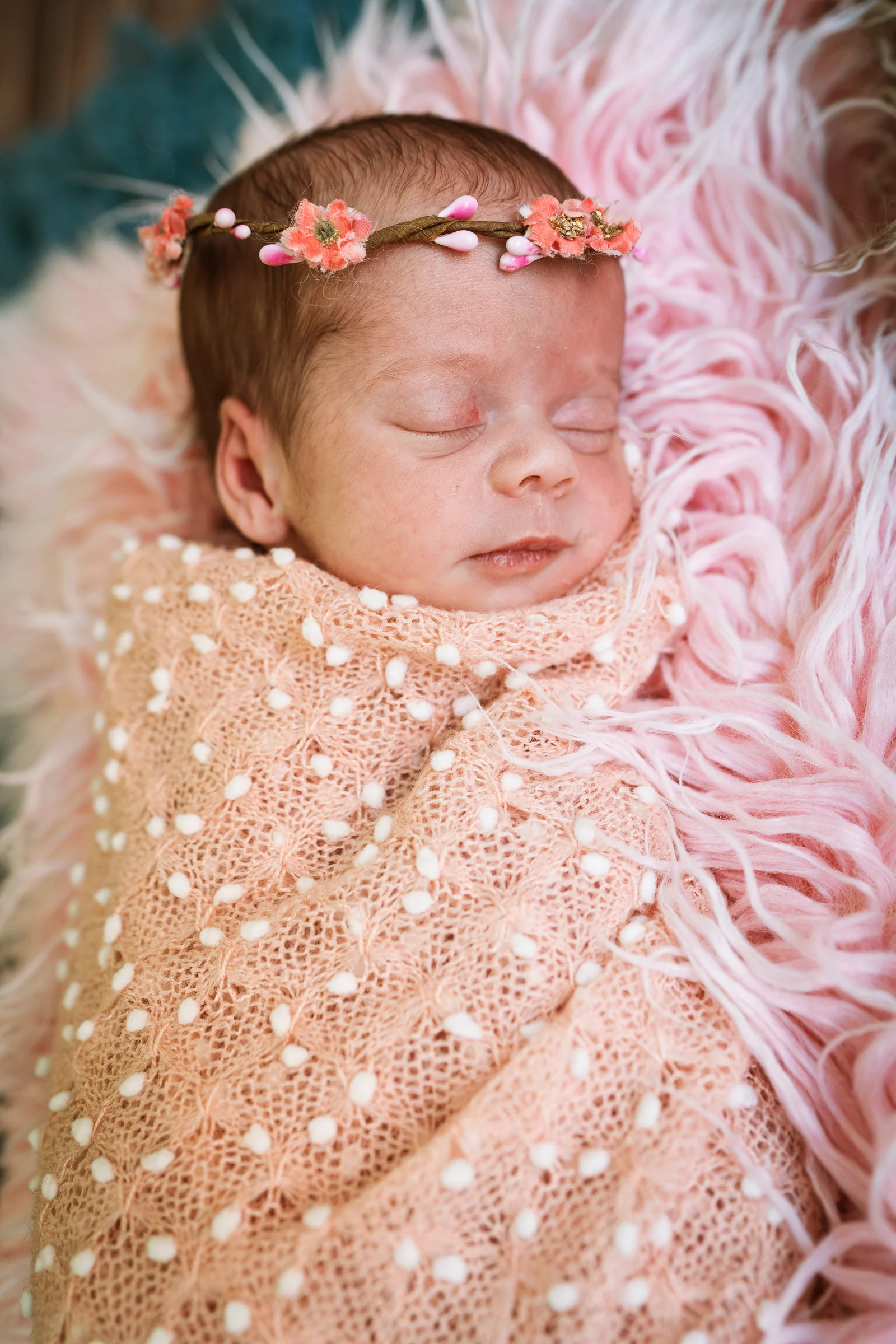 Recién nacida dormida envuelta en tela beige sobre pelo rosa con diadema floral.