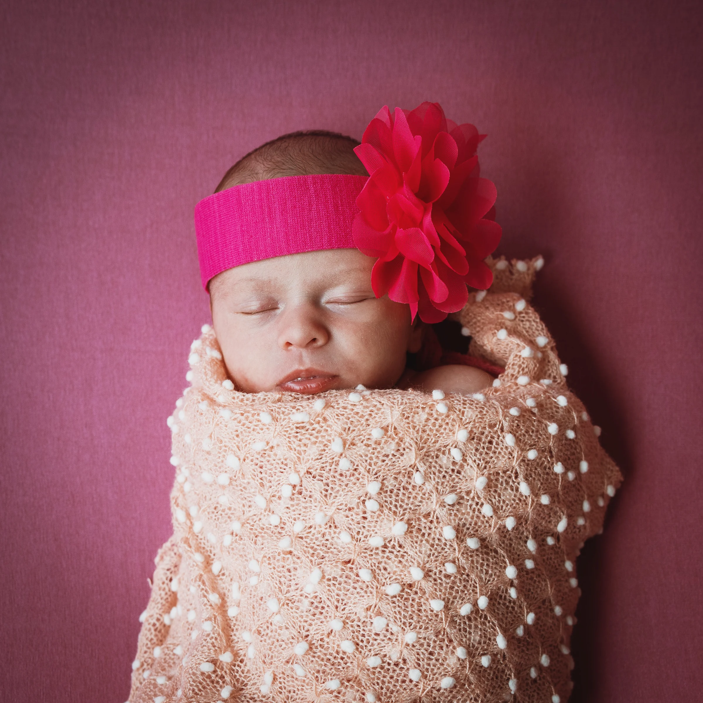 Recién nacida dormida envuelta en tela beige con gran diadema rosa sobre fondo liso.