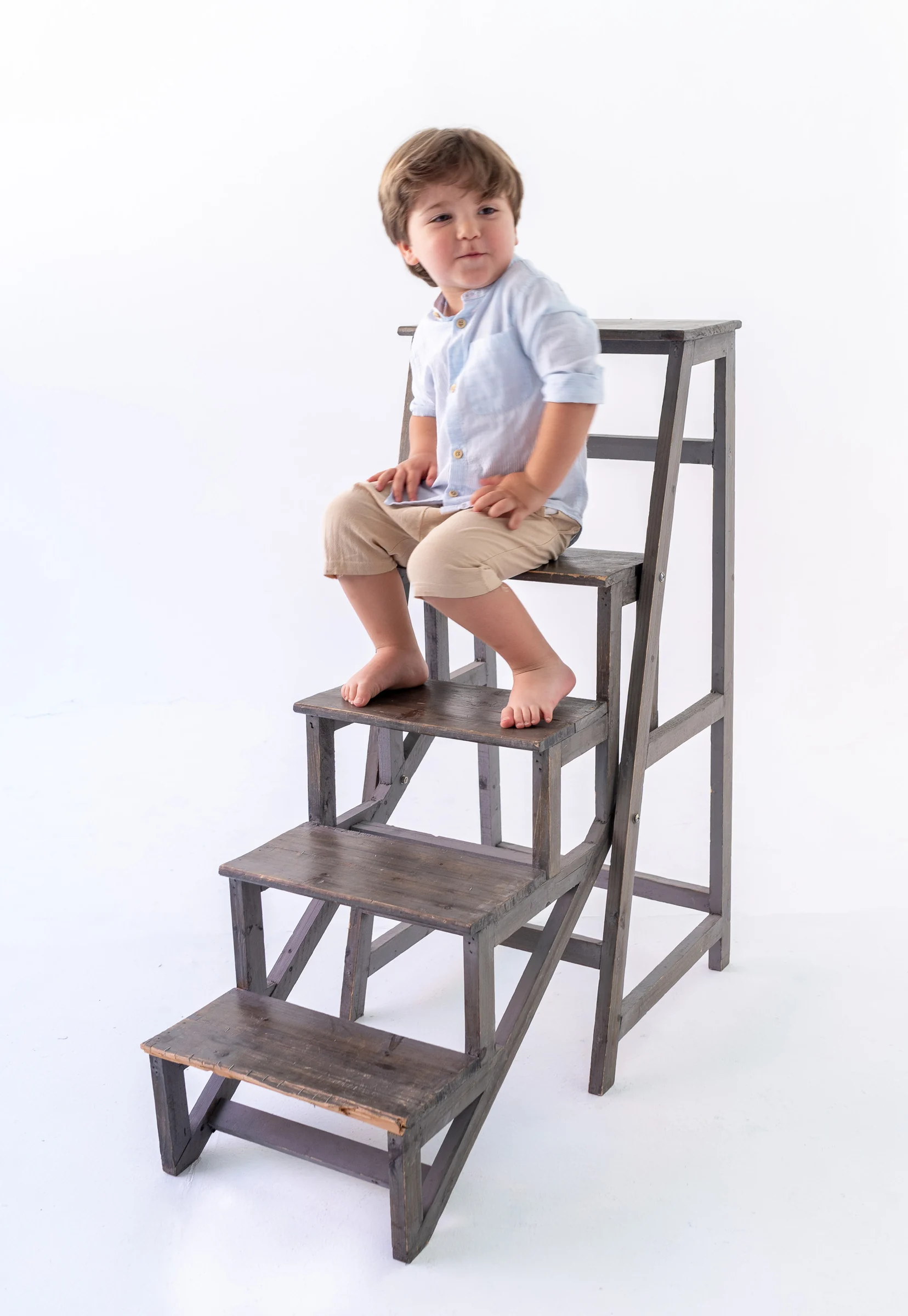 Niño pequeño sentado sobre una escalera de estudio en fondo blanco.