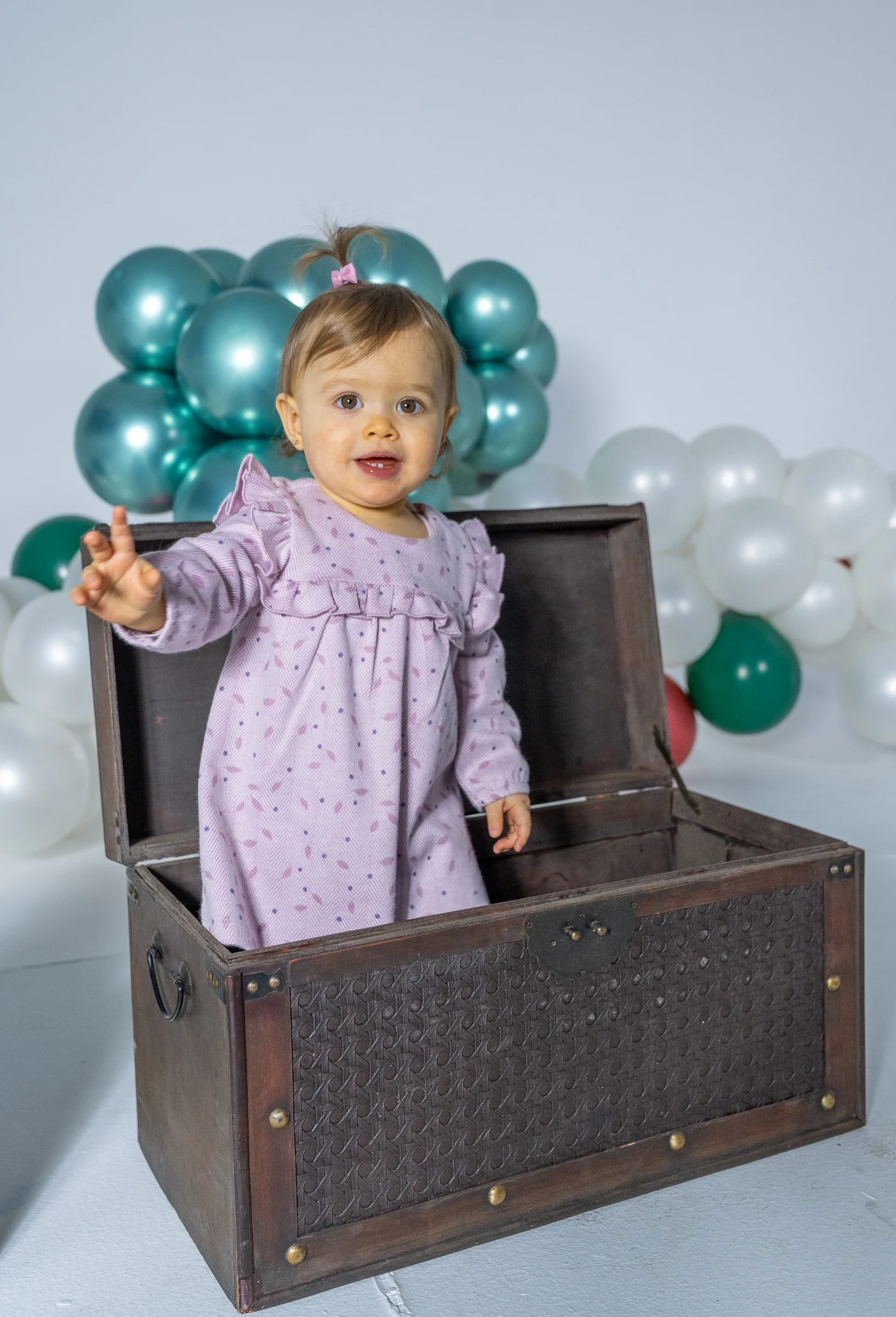 Niña pequeña asomando desde un baúl rodeada de globos en estudio.