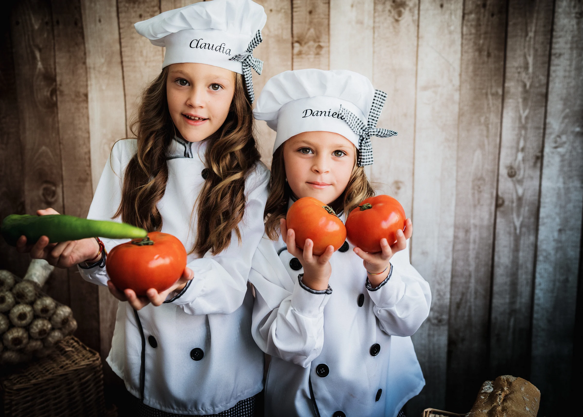 Dos niñas con gorro de cocinera sostienen tomates en un retrato infantil.