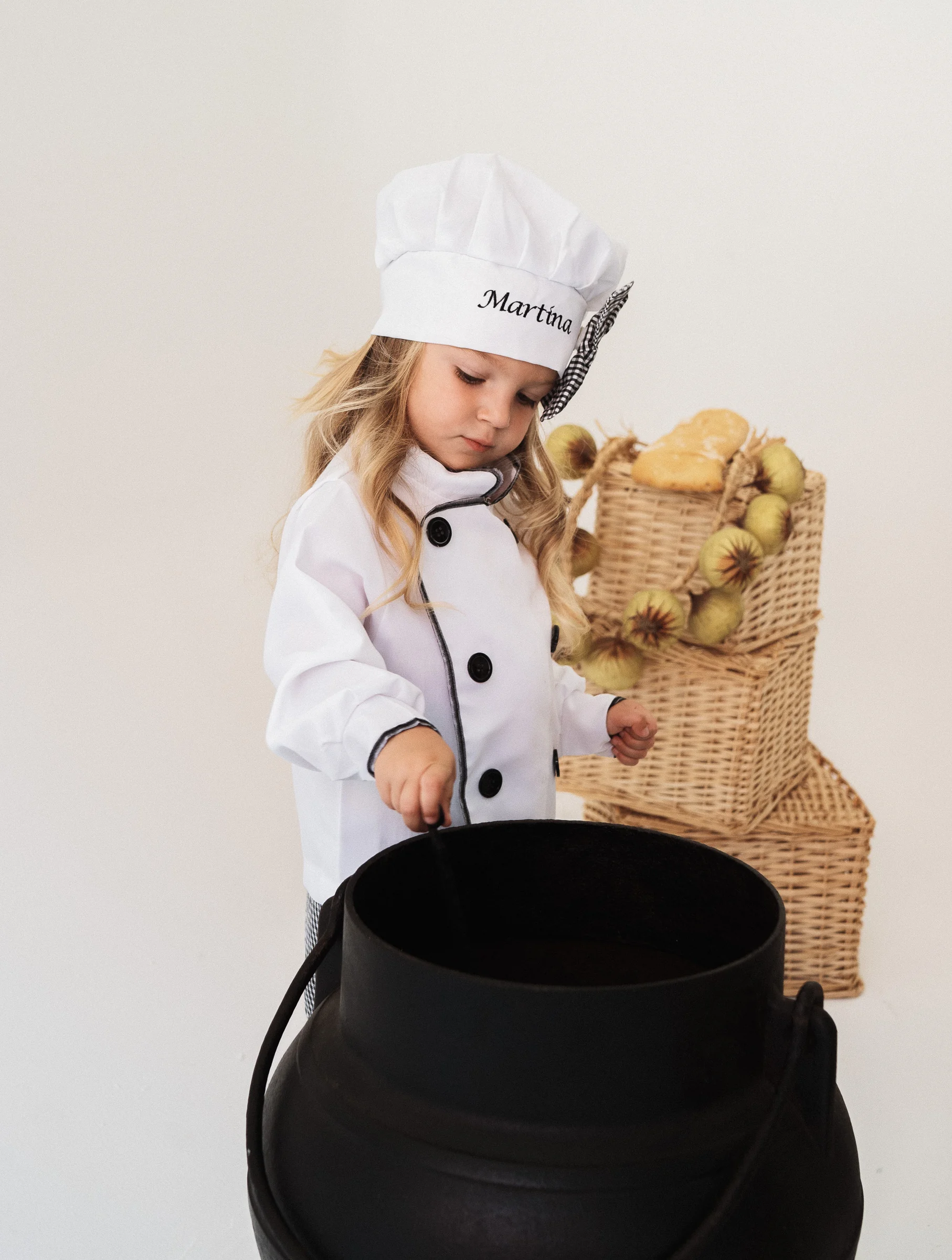 Niña con gorro de cocinera posa junto a una olla en un decorado de estudio.