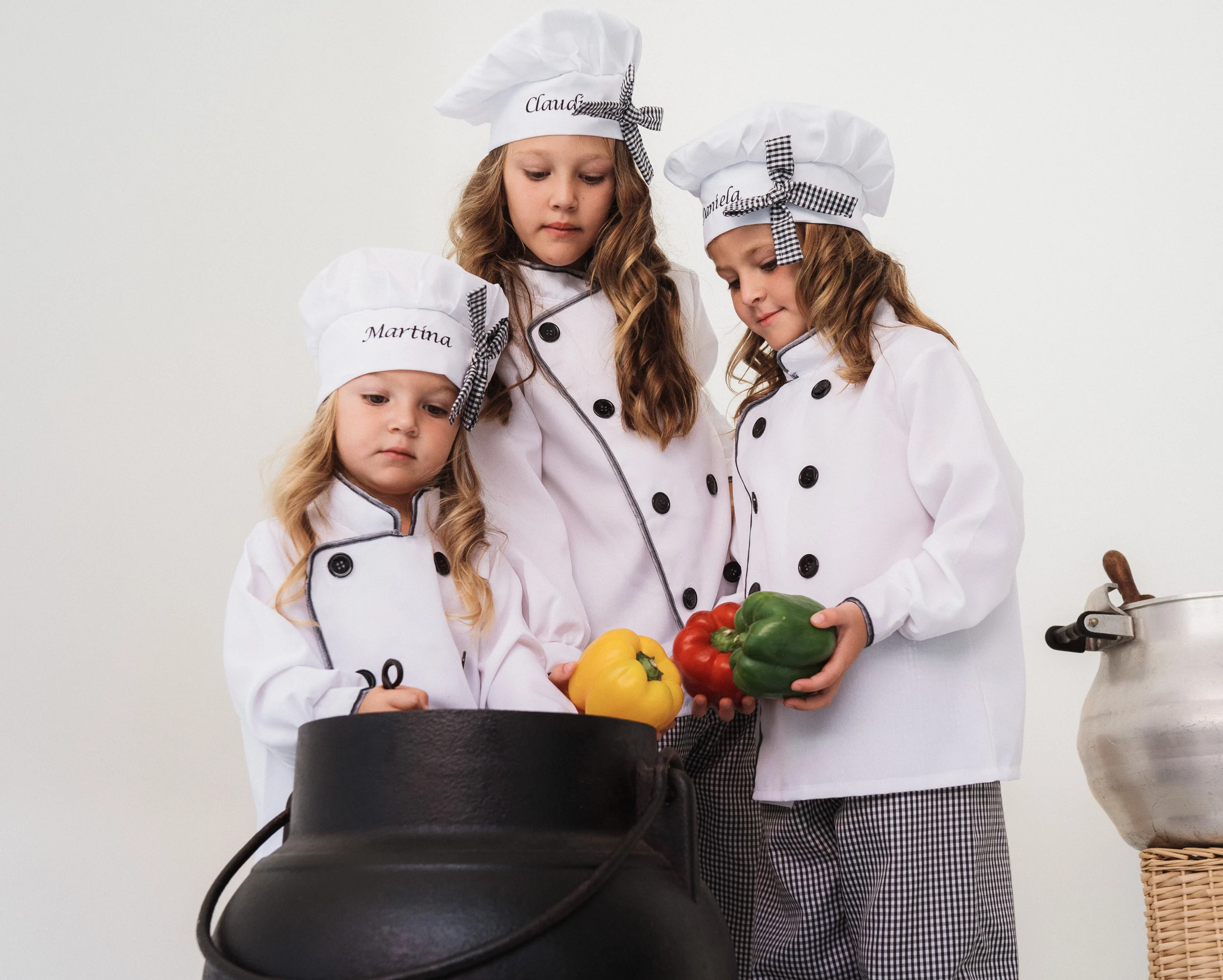 Tres niñas cocineras posan con una olla y cesta de verduras sobre fondo blanco.