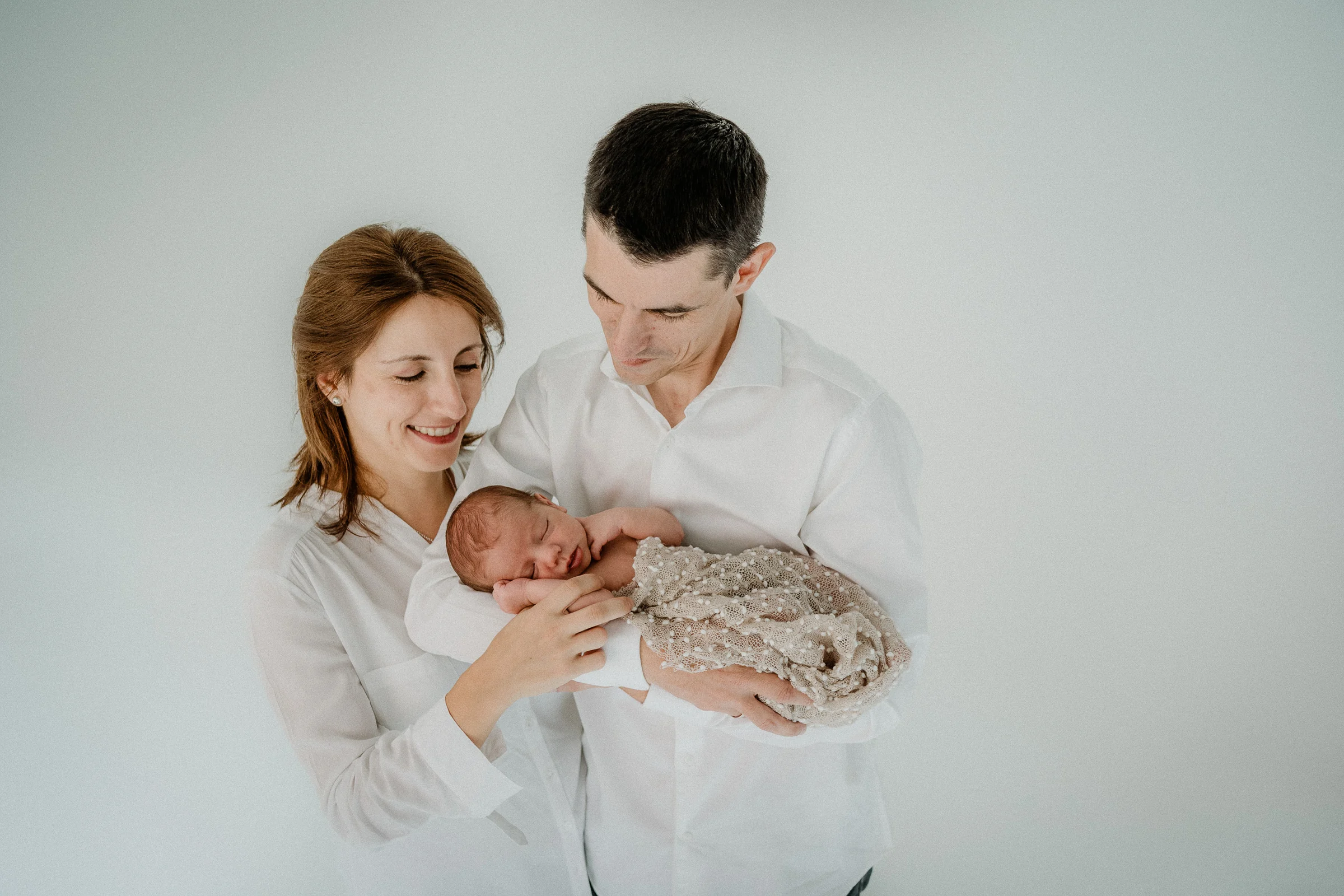 Padres miran con ternura a su recién nacido en brazos en una foto de estudio.