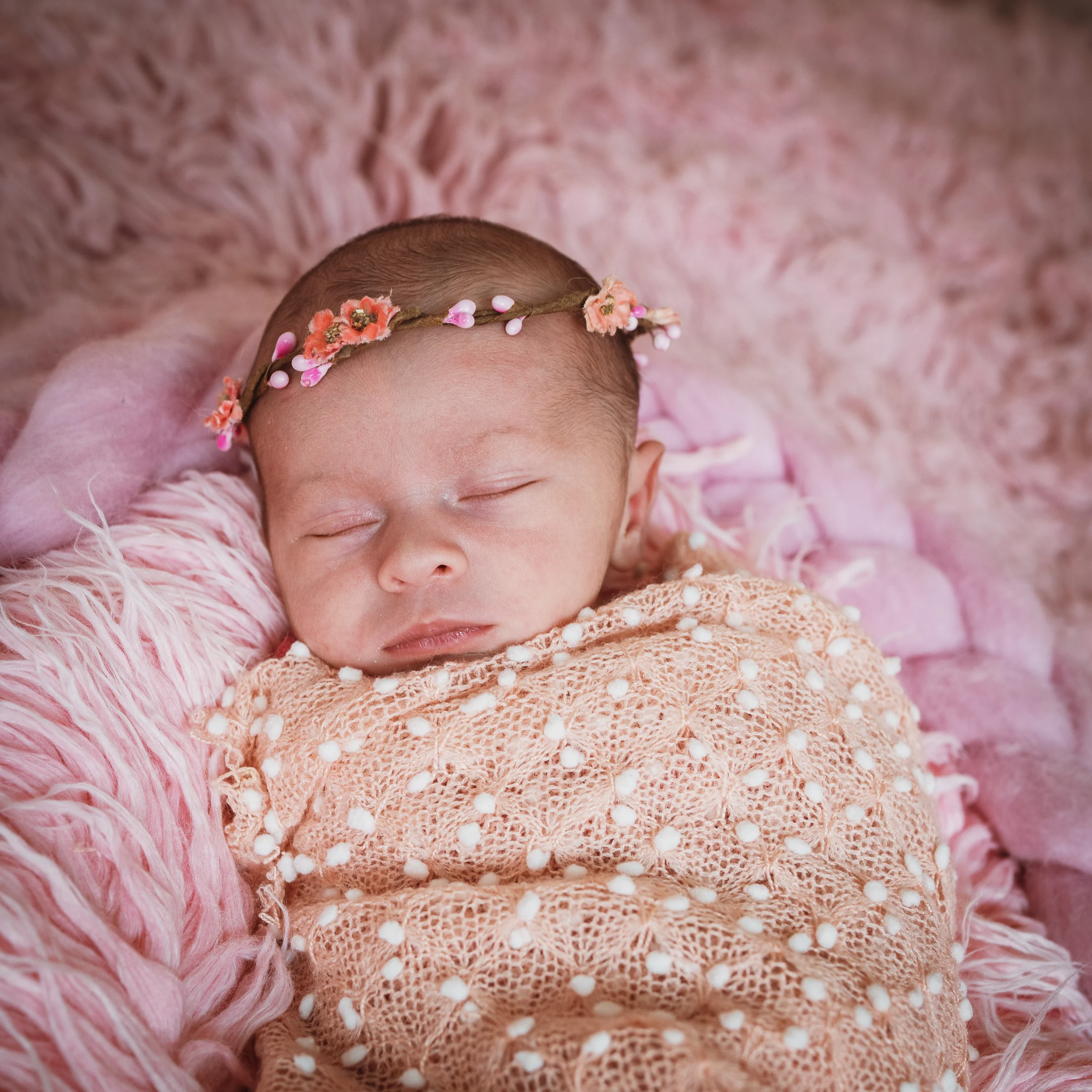 Recién nacida dormida envuelta en manta rosa con diadema floral sobre pelo rosa.