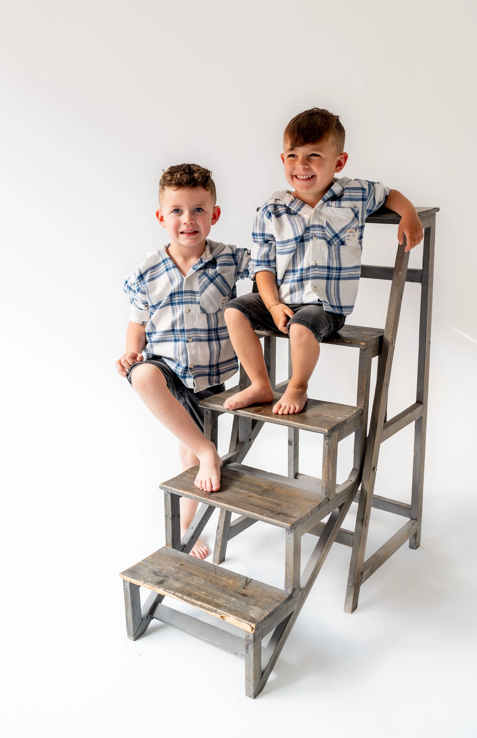 Dos niños posan juntos sobre una escalera de estudio en fondo blanco.