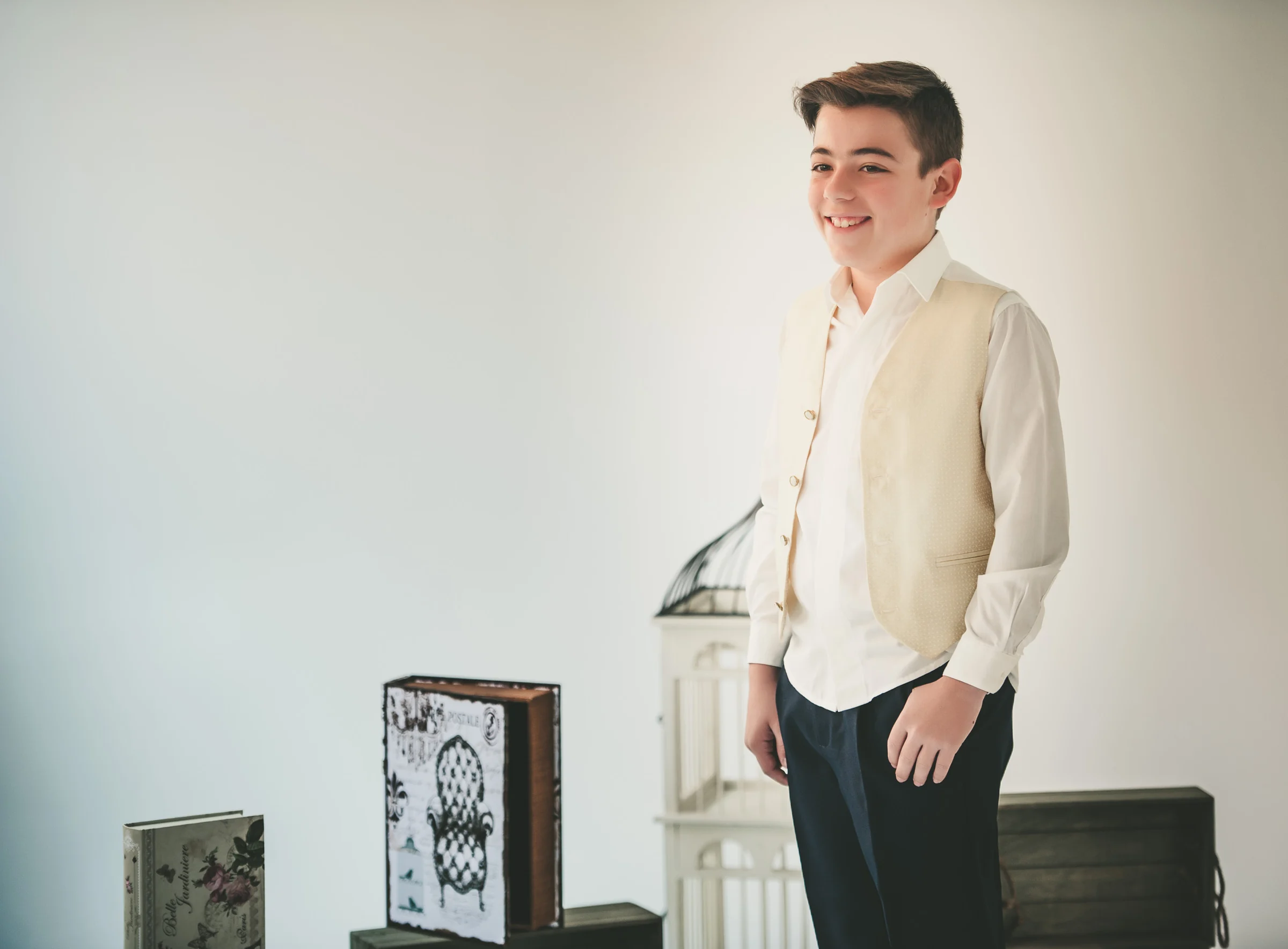 Niño sonríe de pie con camisa blanca y chaleco beige durante una sesión de Primera Comunión en estudio.