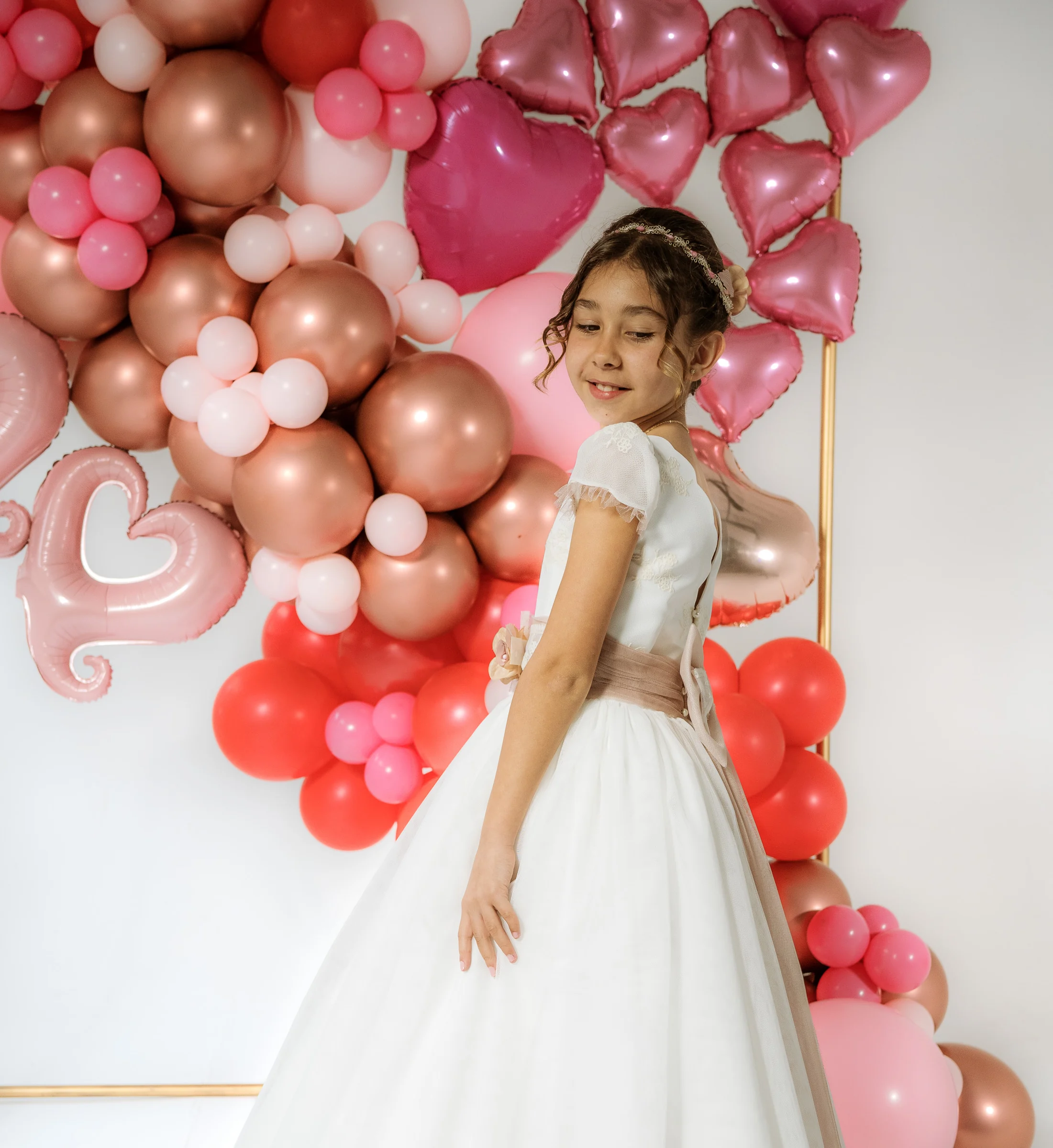 Niña con vestido de Primera Comunión posa de perfil ante una decoración de globos rosas, rojos y dorados.