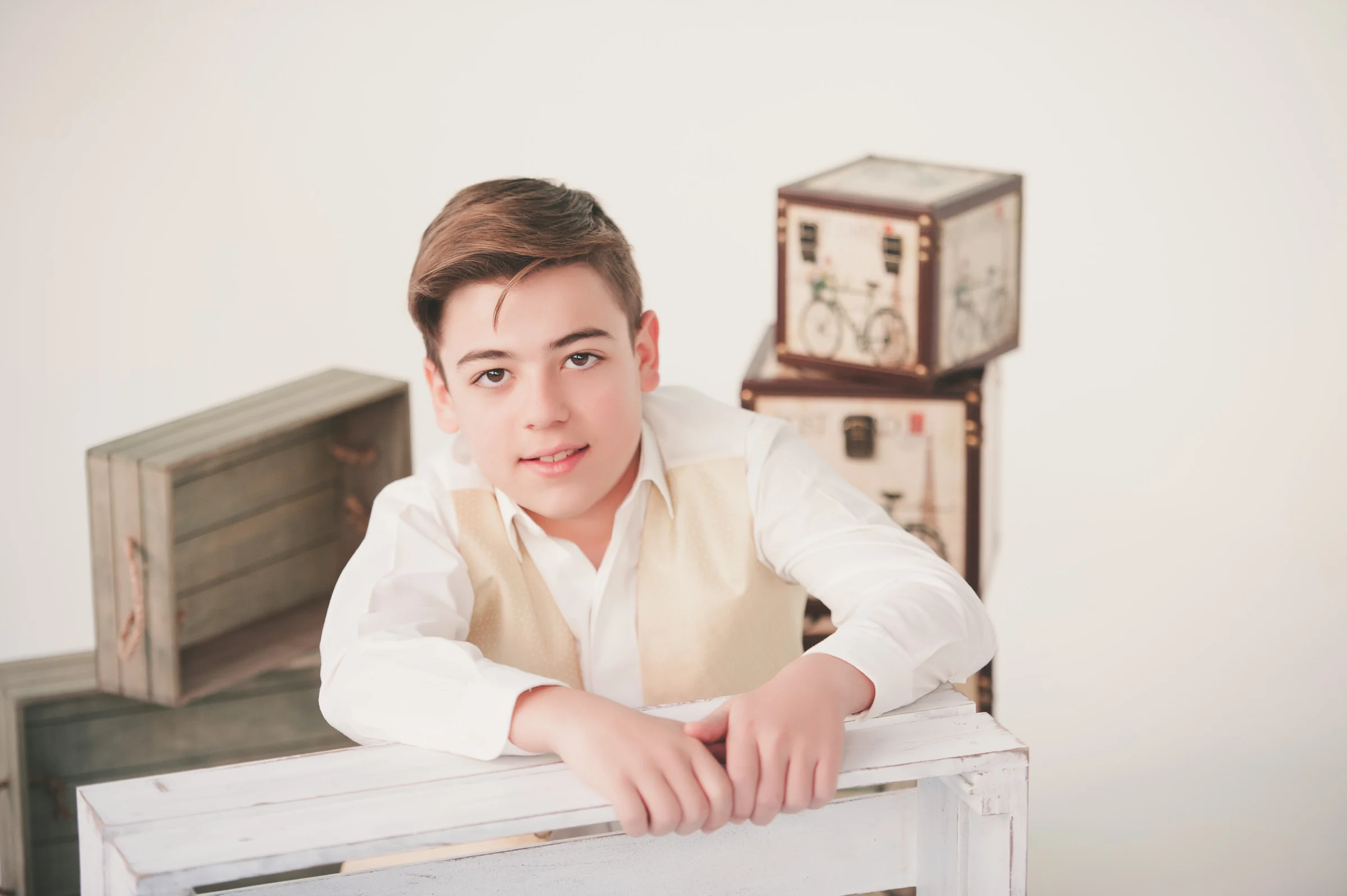Niño con chaleco beige se apoya sobre una caja blanca en un retrato luminoso de Primera Comunión en estudio.