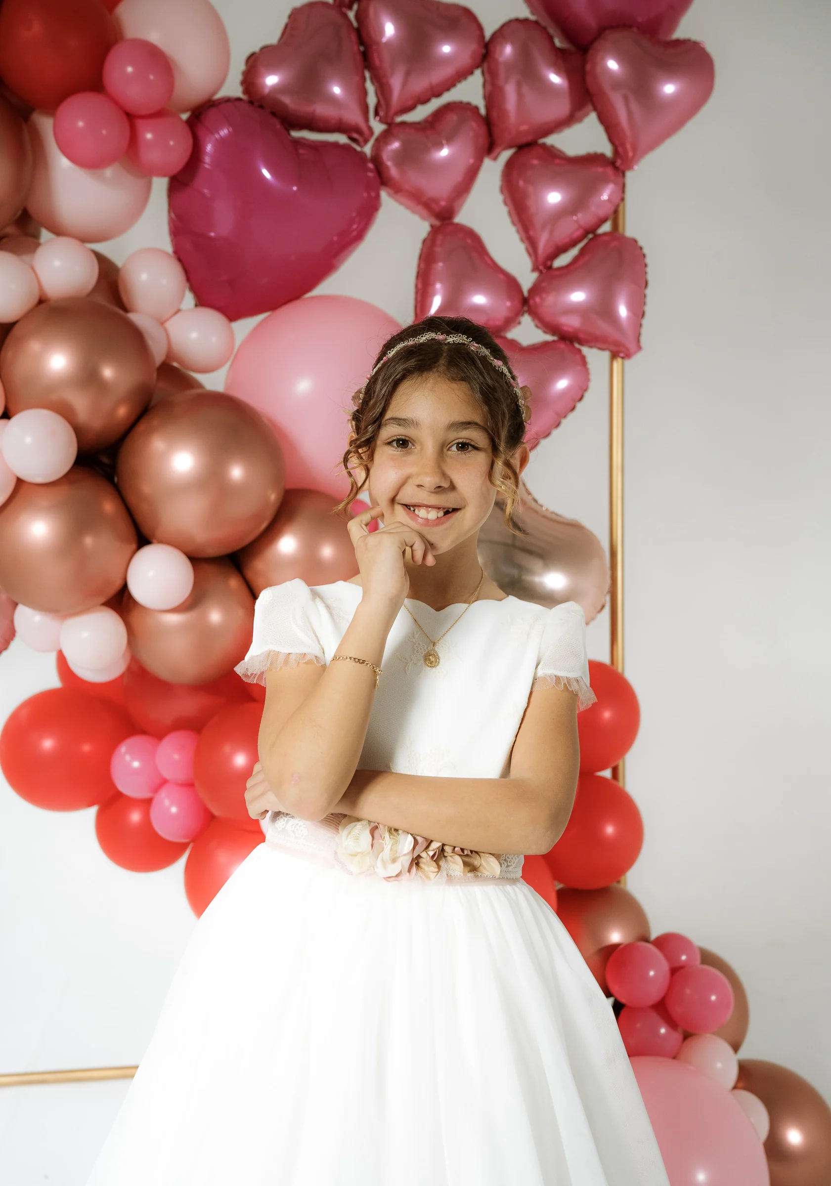 Niña con vestido de Primera Comunión sonríe ante un fondo de globos rosas, rojos y con forma de corazón.