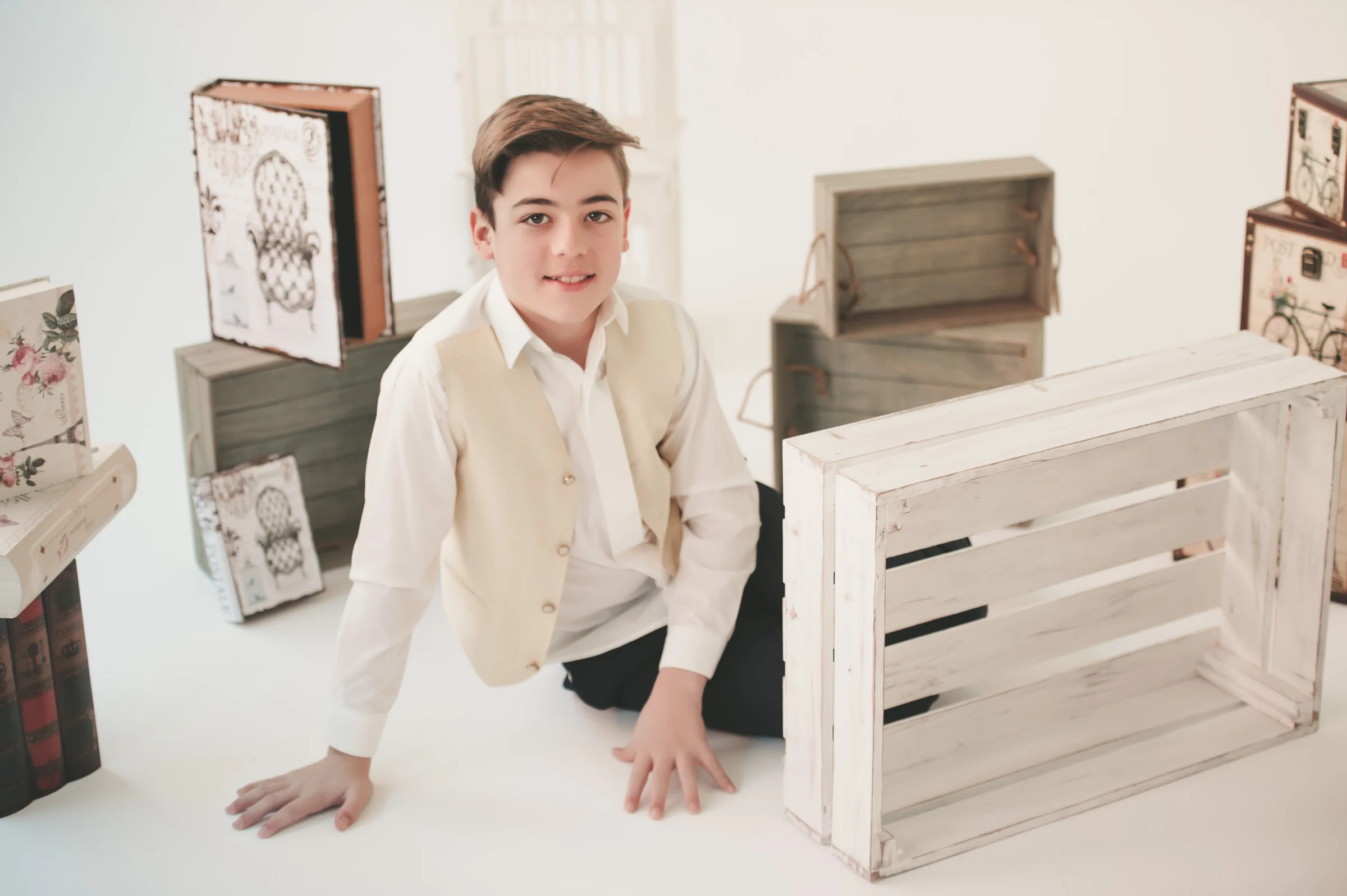 Niño con camisa blanca y chaleco beige posa en el suelo entre cajas y libros decorativos durante su sesión de comunión.