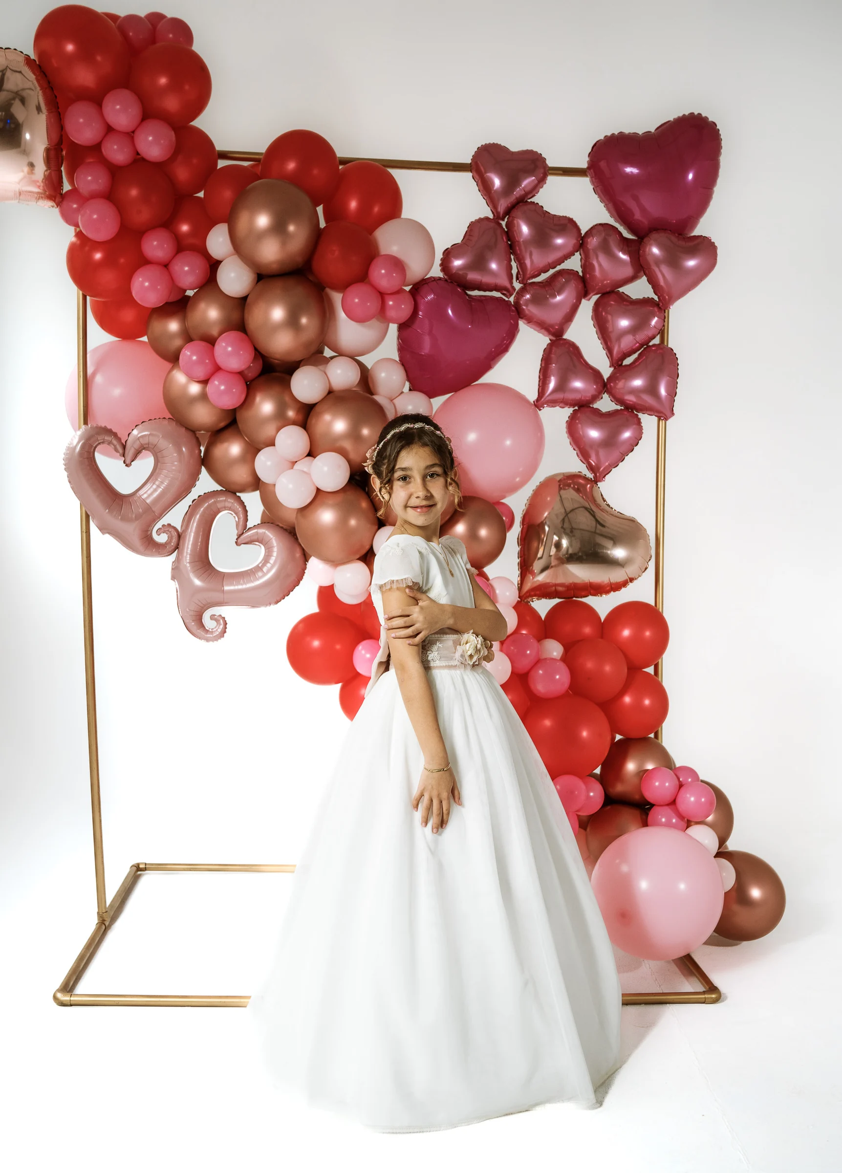 Niña con vestido de Primera Comunión posa de cuerpo entero junto a un arco de globos rosas, rojos y dorados.