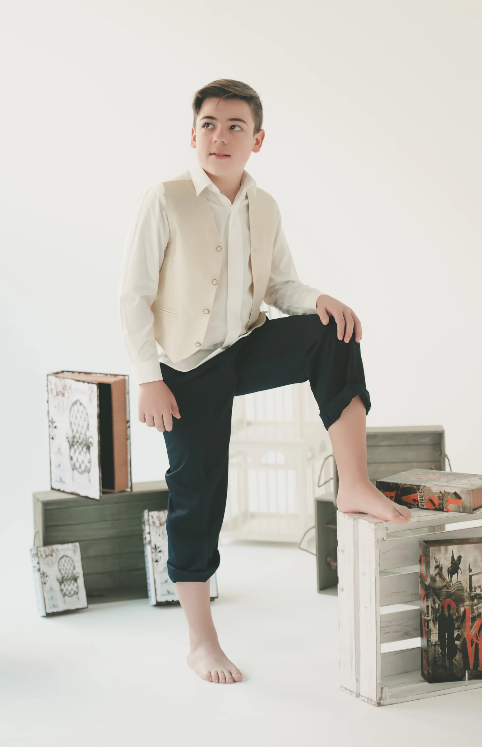 Niño con chaleco beige posa descalzo con un pie sobre una caja blanca en un estudio de Primera Comunión.