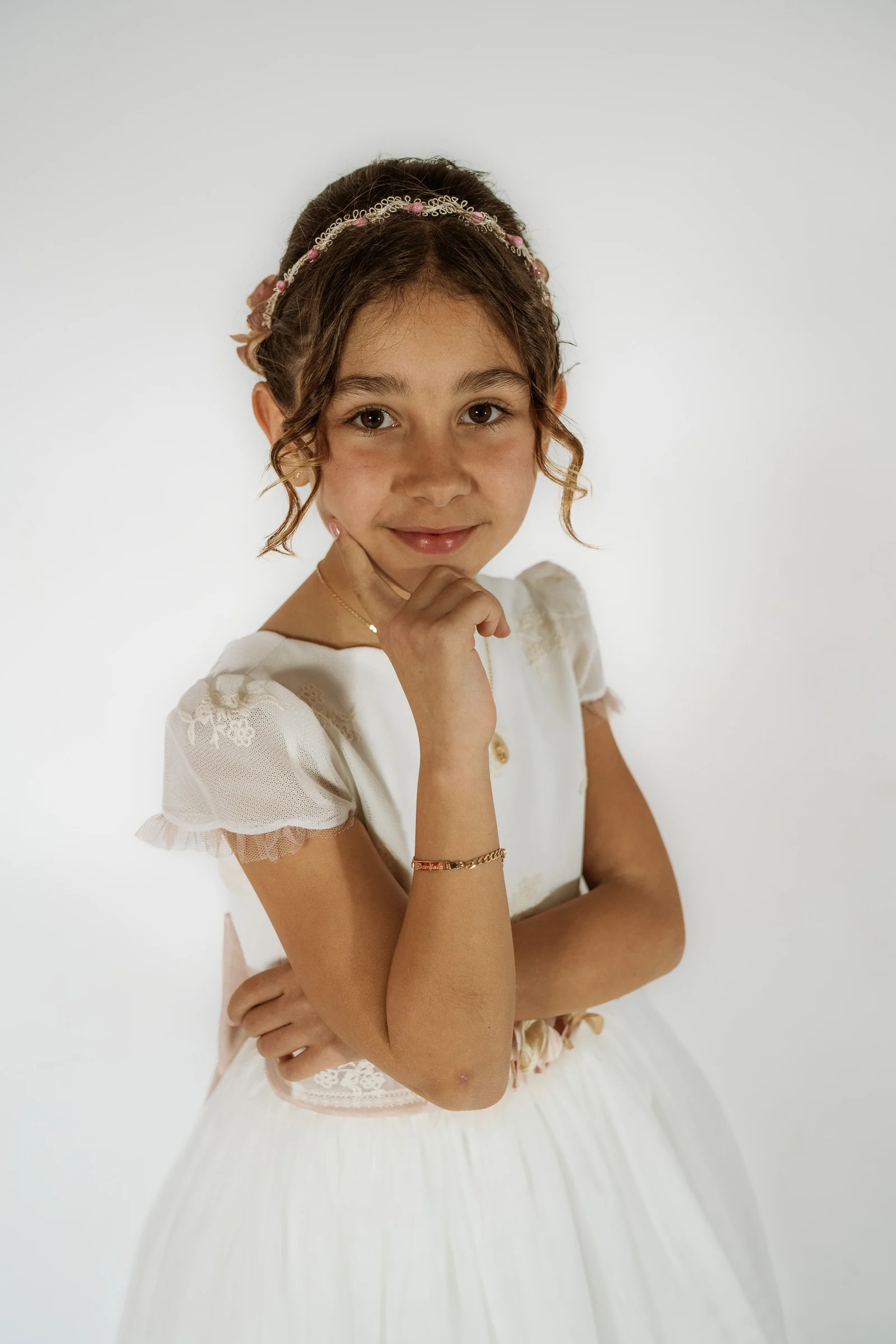 Primer plano de niña con vestido de Primera Comunión y la mano bajo la barbilla sobre un fondo blanco de estudio.